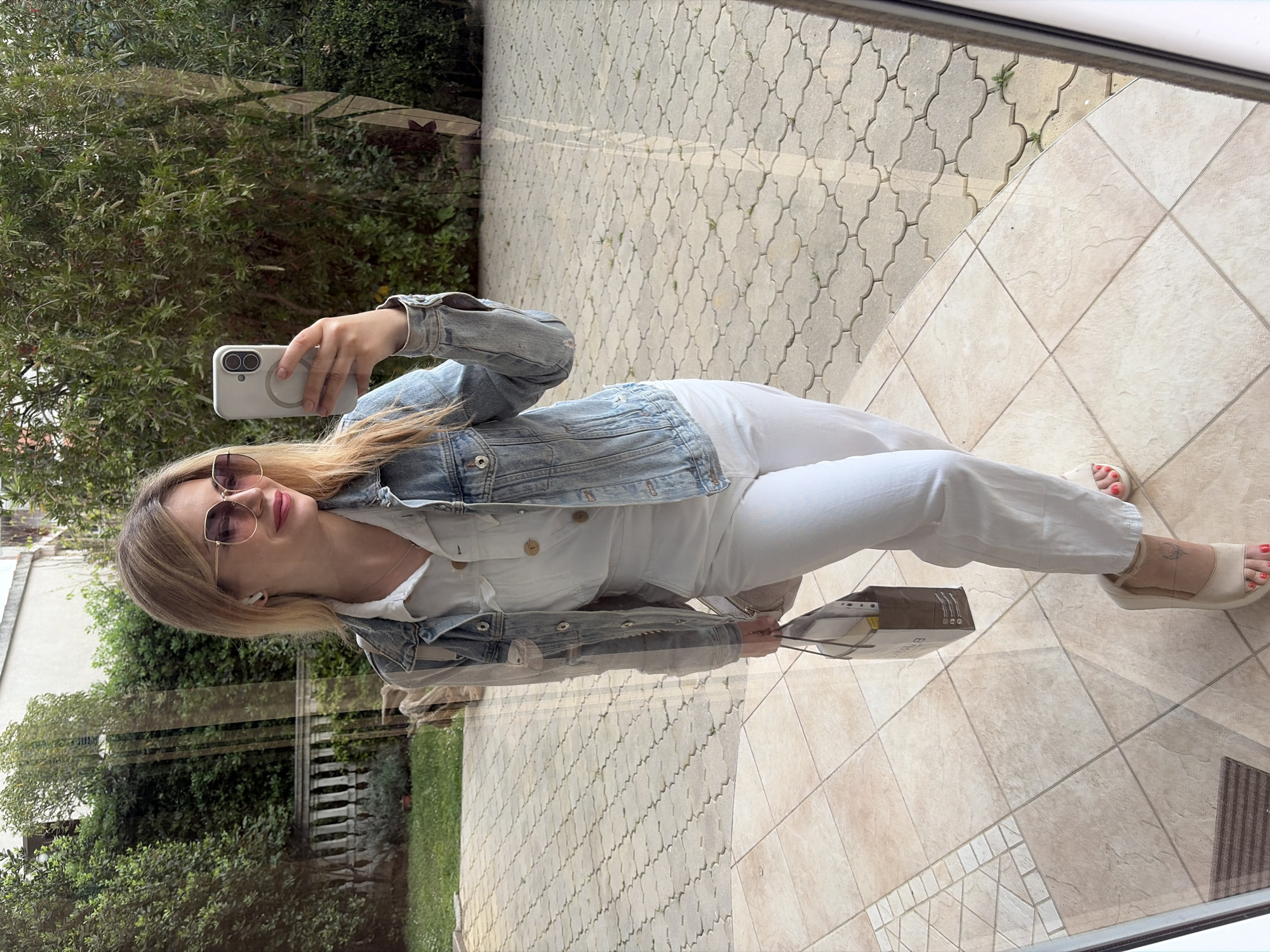 A woman stands inside a building with tile flooring, taking a selfie with a smartphone. She is wearing sunglasses, a denim jacket, and white pants. A shopping bag rests on the floor near her feet. The scene is reflected in a glass panel, showing a garden and a walkway paved with decorative stone. The location is in the city of Budva, Montenegro.