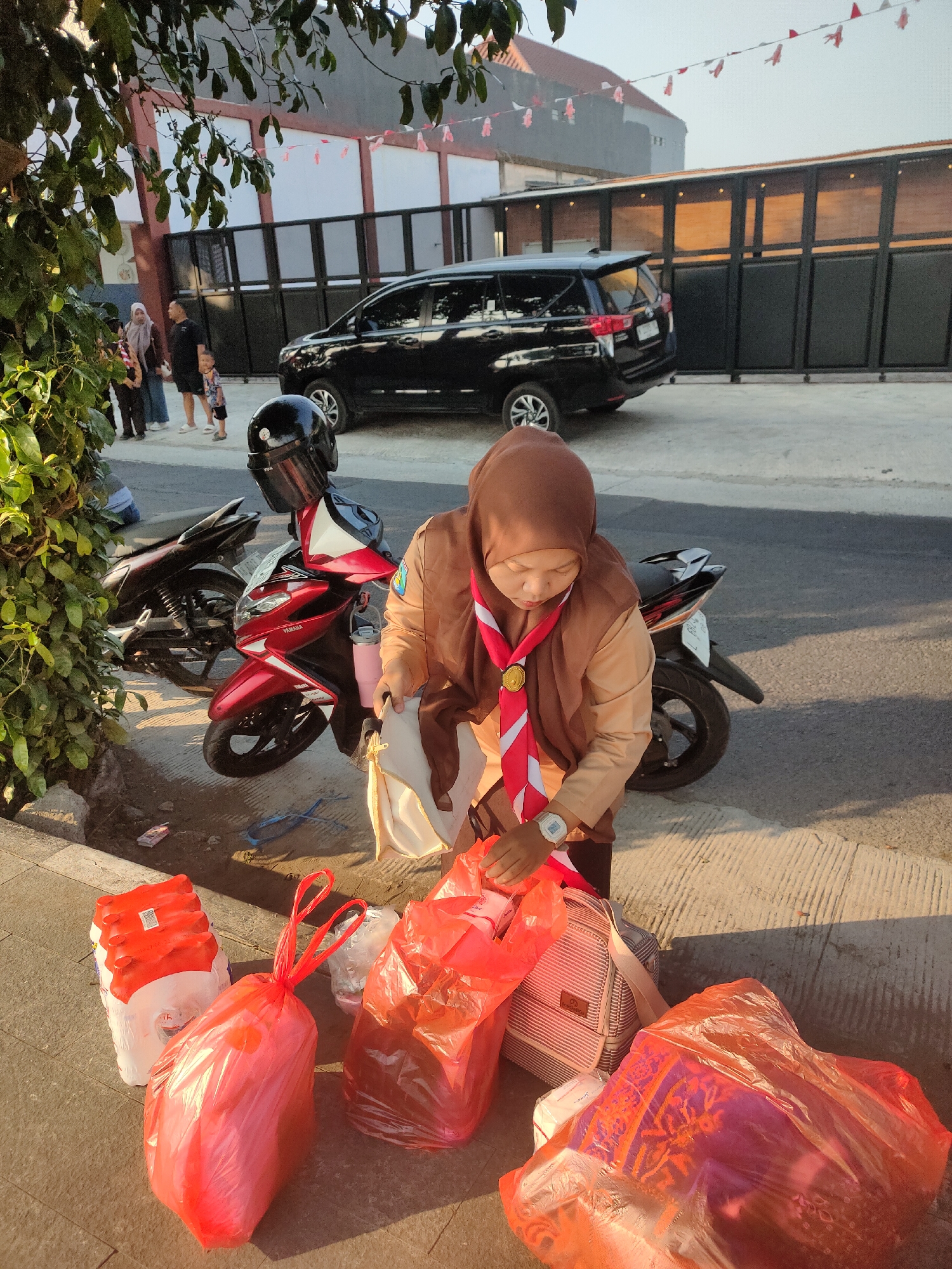 A woman in a brown hijab and a light brown uniform with a red and white sash is standing next to a red motorcycle on a street in Ngasem, Indonesia. She is reaching into a red plastic bag, holding a white bag in her other hand. In front of her, on the pavement, are several other red plastic bags, a stack of red and white packaged items, and a striped bag. Behind her, another motorcycle is parked. In the background, a black van is visible, and a group of people are standing near a building with black gates. Red and white flags are strung across the top of the frame. The sun is casting long shadows, indicating it is likely late afternoon or early morning.