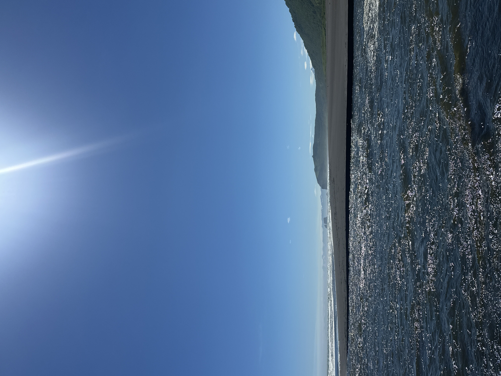 The image shows a wide, dark-water river flowing towards a black sand beach and the ocean under a clear, bright blue sky with a visible sun flare in the upper portion. The river in the foreground is choppy with sunlight reflecting off the surface. Beyond the river, the black sand beach stretches horizontally, meeting a calm ocean with gentle waves. In the background, a lush green, forested mountain rises from the right side of the frame, and a rocky promontory juts out into the sea on the left. The scene is devoid of people or any man-made structures. This location is likely on the coast of Petropavlovsk-Kamchatskiy, Russia, known for its volcanic landscapes and black sand beaches, such as Khalaktyrsky Beach.