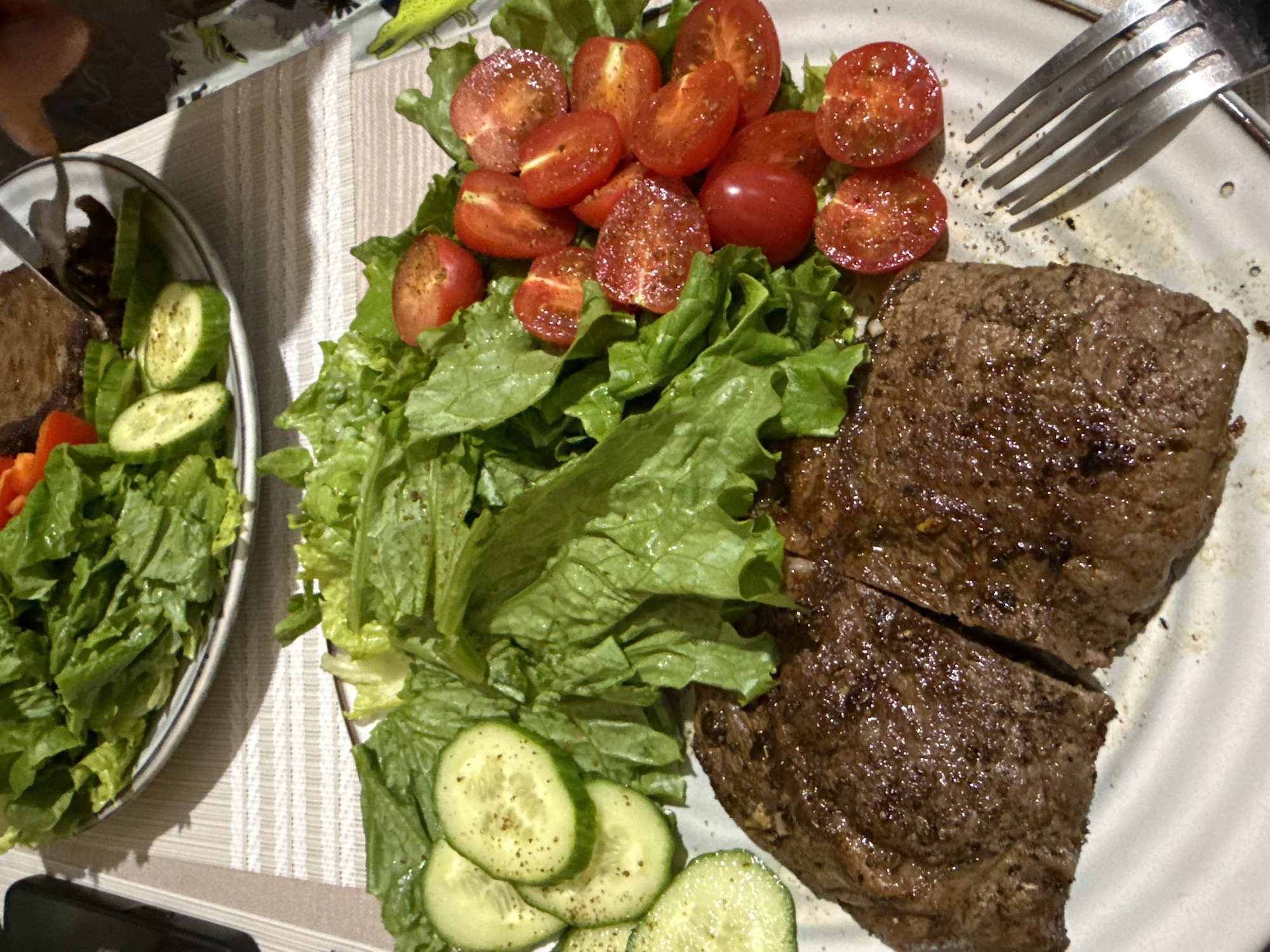 The image shows two plates of food on a table with a striped tablecloth.
Plate 1 (right): Contains a sliced steak, lettuce, halved cherry tomatoes, and cucumber slices. A fork is on the upper right.
Plate 2 (left): Contains a similar salad with lettuce, cucumber, and a red bell pepper slice. There is a dark colored protein.
The location is not discernable. There is no information about subjects, objects, interactions, or environment.