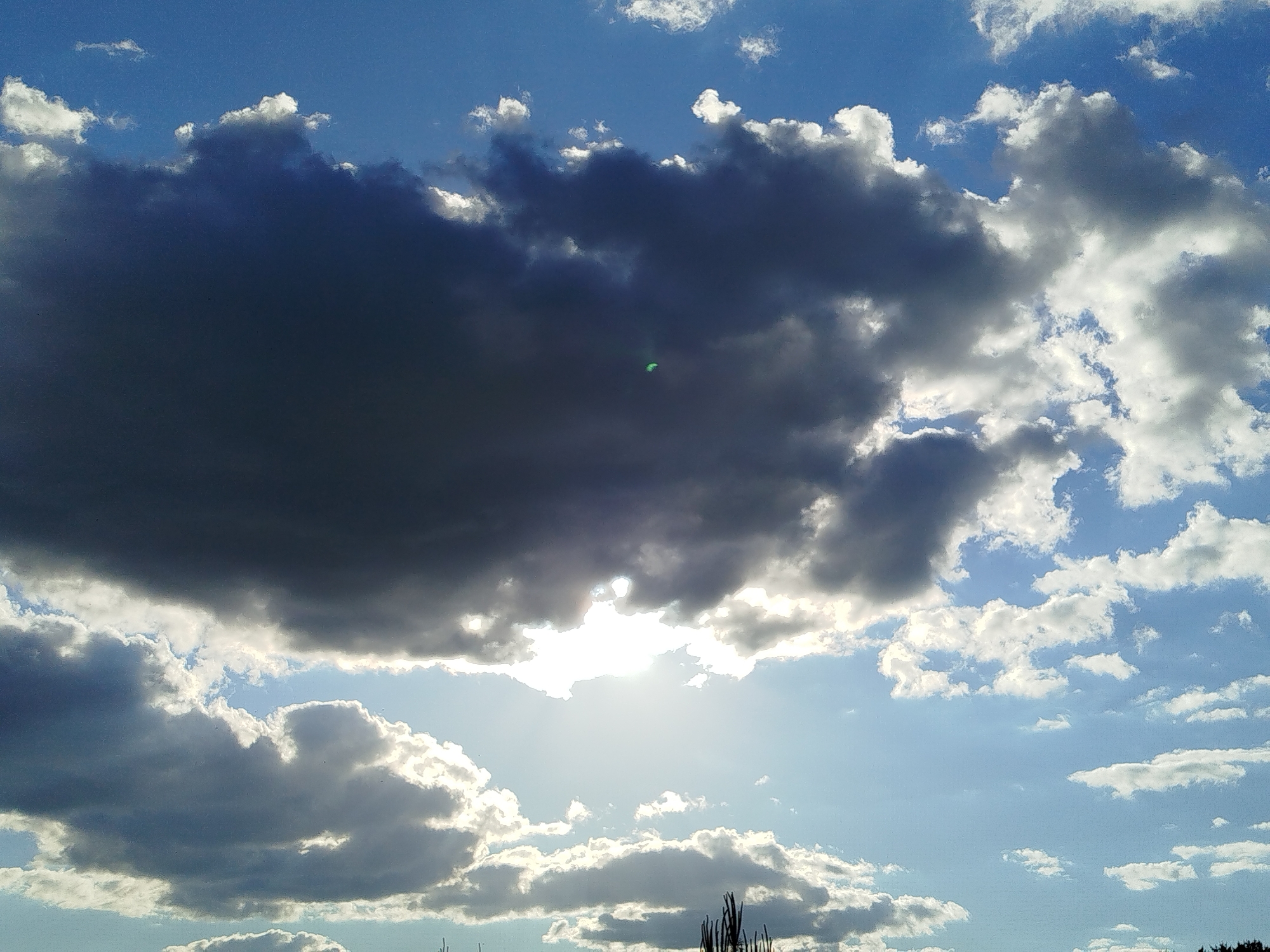 The image shows a bright, sunny day with scattered clouds. A large, dark cloud dominates the upper left portion of the frame, while smaller, white clouds are spread throughout the rest of the sky. The sun is partially obscured by the clouds, creating a bright, diffused light. In the lower part of the image, what appears to be tree tops can be seen. The location is identified as Заречье, Russia.