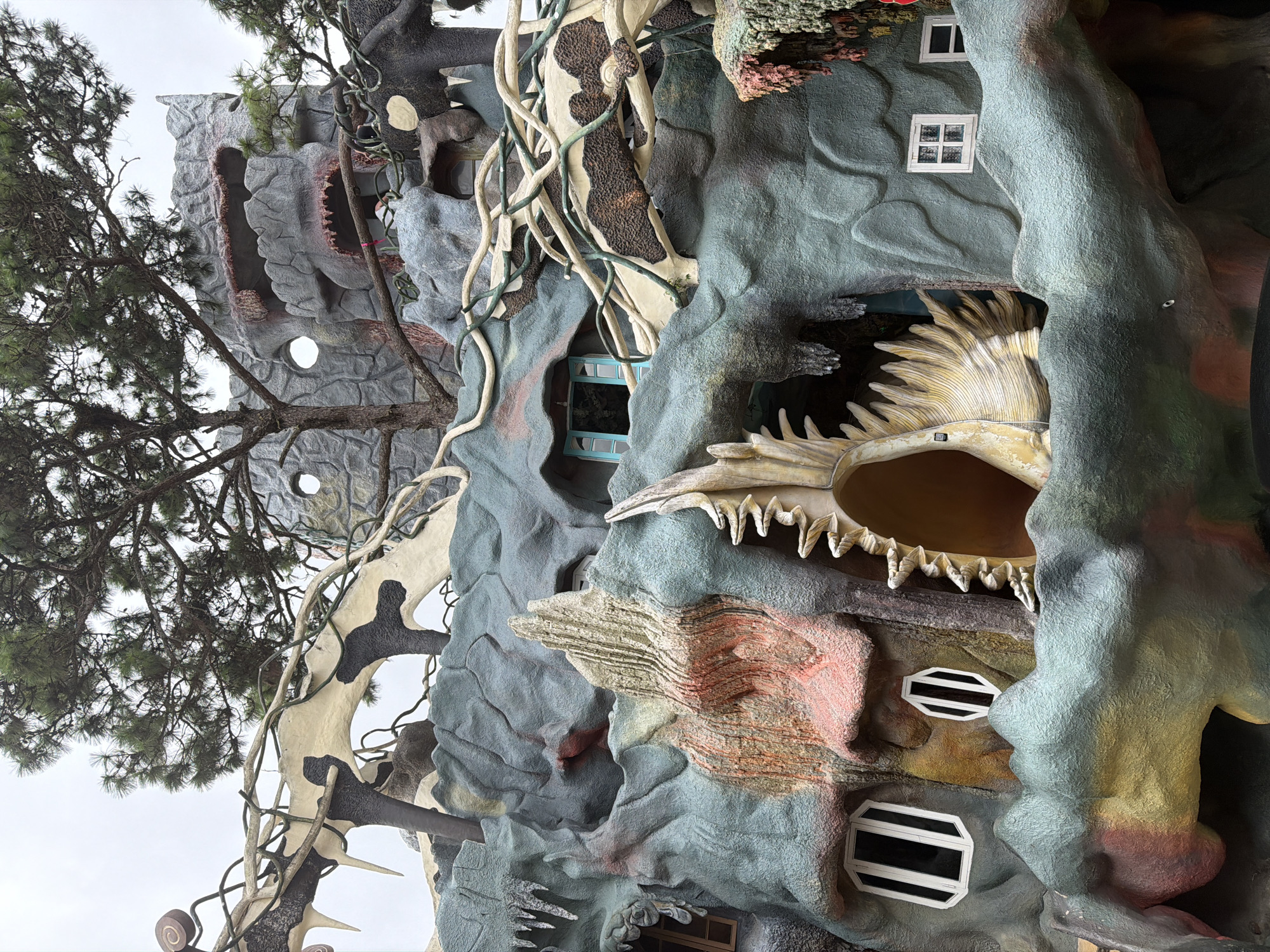 This is a photograph of the exterior of the Crazy House (Hang Nga Guesthouse) in Da Lat, Vietnam. The building's architecture is organic and surreal, resembling a natural landscape with cave-like structures, winding vines, and carved figures. The facade is textured and painted in muted, earthy tones with accents of red, orange, and green. Several windows of various shapes and sizes are visible, some with blue shutters. A large, shell-like opening with sharp, tooth-like protrusions dominates the center of the image, appearing like a gaping mouth. A large tree with pine needles branches out from the top of the structure, its branches intertwined with the building's design. The sky is overcast, suggesting a cloudy day. There are no people visible in the image.