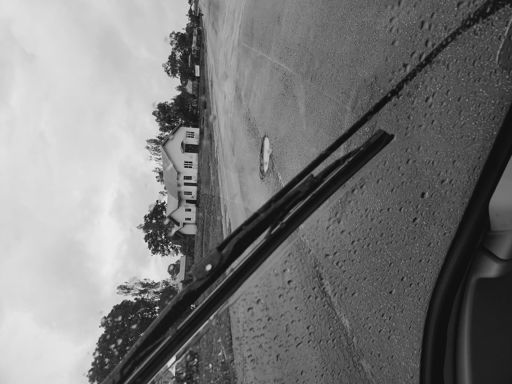 The black and white photograph presents a view from inside a vehicle, looking out through a rain-splashed windshield. Numerous water droplets cling to the glass, suggesting recent or ongoing rain. Two windshield wiper blades are prominently featured in the foreground, resting horizontally at the bottom of the frame, slightly angled upwards to the left. A portion of the vehicle's interior, possibly the right door frame or dashboard, is visible along the very right edge.
Beyond the wet glass, a wide, wet asphalt road or parking lot stretches across the mid-ground, showing reflective patches from the water. A distinct, light-colored puddle or pothole filled with water is visible on the road surface. In the background, on the left side of the scene, stands a simple, light-colored building, possibly a two-story house or institutional structure, characterized by several rectangular windows. Lush, dark trees with dense foliage are clustered around and behind the building. The sky above is overcast and bright, indicative of a cloudy day.
The scene captures a moment during or shortly after rain, evoking a quiet or subdued atmosphere. The time of day appears to be daytime, though the heavy clouds obscure any direct sunlight. No people are visible, and there is no discernible text in the image. The setting suggests a semi-urban or rural environment, consistent with the provided location context of Vom, Nigeria.