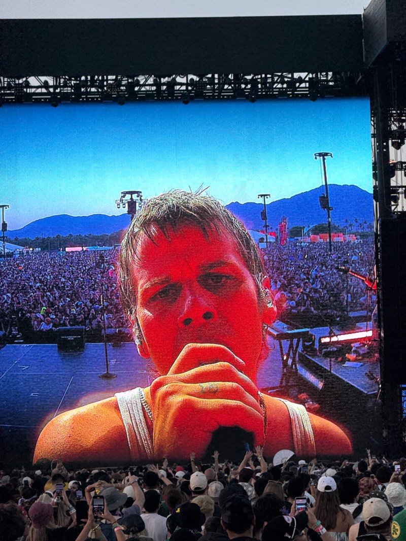 A large crowd of people at the Coachella music festival in Indio, California, are watching a close-up image of a singer on a giant screen. The singer is holding a microphone and appears to be singing. The background shows a stage, more of the crowd, and distant mountains under a blue sky. Metal infrastructure is above the screen. The crowd is mostly standing, with some people holding up cell phones, possibly recording the performance.