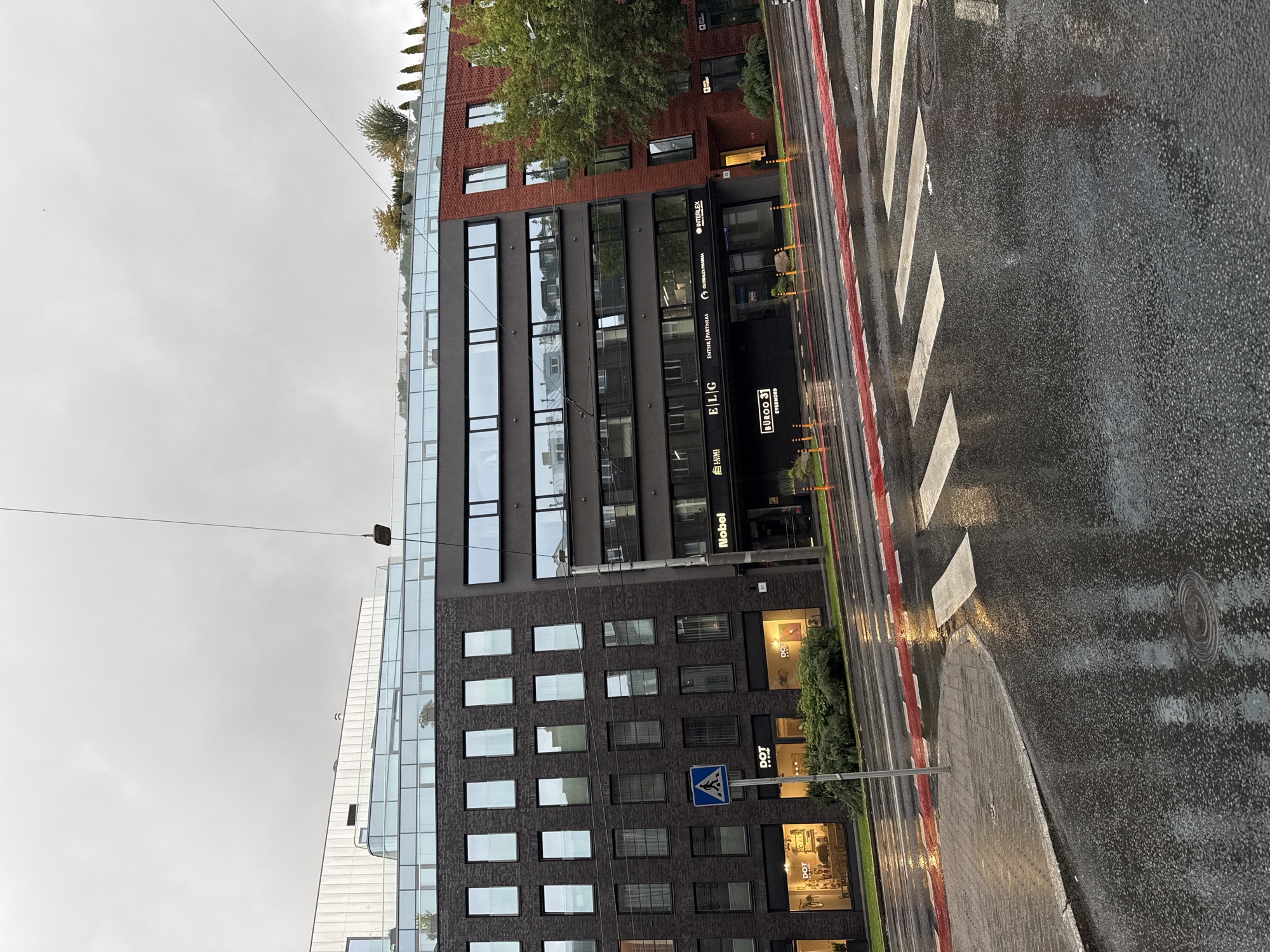 The image depicts a modern, multi-story building in Tallinn, Estonia, on a cloudy day with rain evident on the wet street and sidewalk. The building features a dark brick facade on the left, transitioning to a dark glass facade in the middle, and a red brick facade on the right. Several businesses have signage on the ground floor, including "DOT", "Nobel", and "BUÜRO 31 EVERNHORD", among others. A pedestrian crossing is visible on the wet road in the foreground, marked by white stripes and a red border. A street light hangs from a wire above the scene. There are no people visible in the image. The environment appears urban, with traffic infrastructure like a street and sidewalk present.
