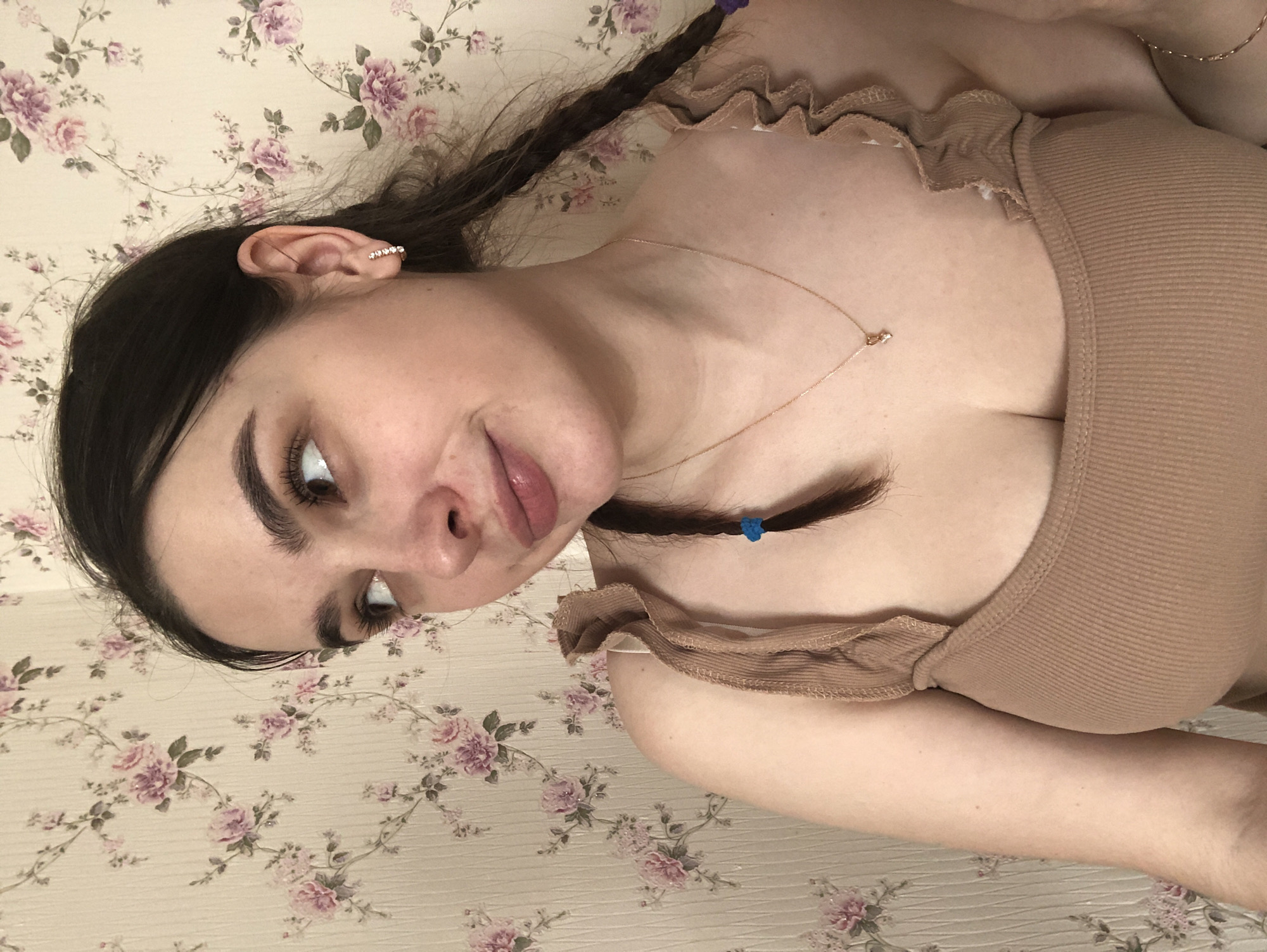 The image displays a close-up portrait of a female subject, visible from the mid-chest upwards, situated indoors. The subject is a young adult with dark hair, styled in at least two braids, one visible over her right shoulder secured with a blue elastic, and another partially seen on her left with a purple elastic. Her skin tone is fair. She wears a light brown, ribbed tank top featuring ruffled straps. Accessories include a gold-toned hoop earring with multiple small clear stones in her right earlobe, a thin gold-toned necklace with a small, unidentifiable pendant, and a thin gold-toned bracelet partially visible on her left wrist. Her eyebrows are dark and full, and her eyelashes appear long. She is looking upwards and to her right, away from the camera, with a slight, subtle smile or smirk on her tinted lips. The background is a wall covered in light beige wallpaper patterned with repeating clusters of pink and purple roses and green leaves. The specific city of Noviy, Russia, cannot be determined from the visual information present in the image.