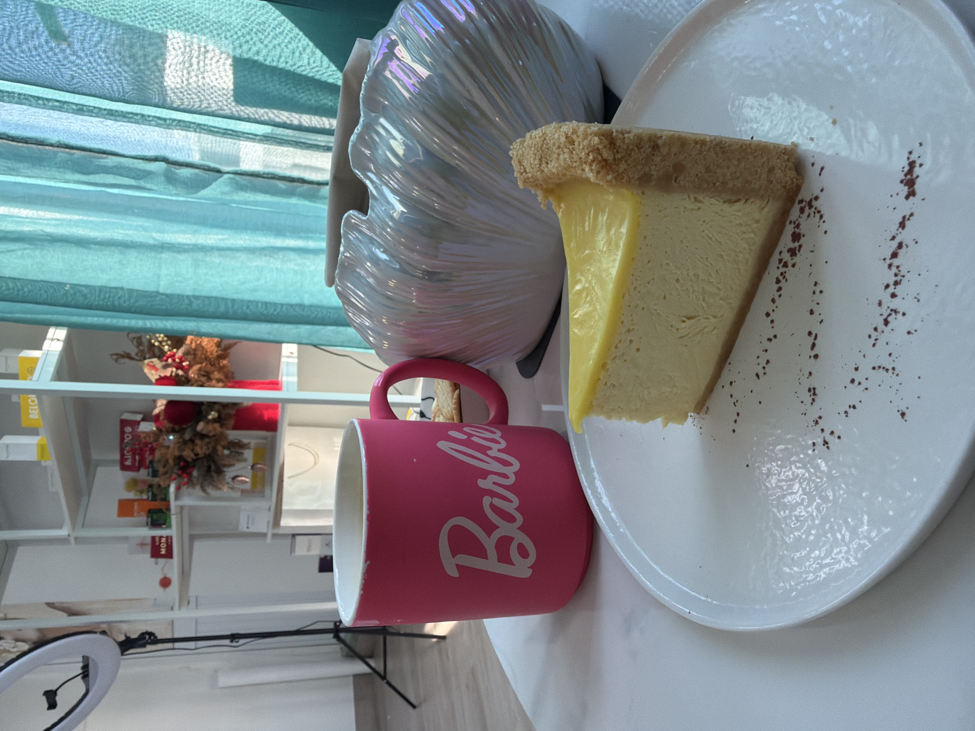 The image depicts a close-up, eye-level view of a slice of cheesecake on a white plate, placed on a white table. The cheesecake has a graham cracker crust and a smooth, pale yellow filling topped with a thin layer of glossy yellow topping. Dark brown powder, possibly cocoa or cinnamon, is sprinkled in small dots and a line on the plate. To the left of the plate is a bright pink mug with "Barbie" written in white script. The mug appears to be filled with a dark liquid, presumably coffee. Behind the mug, to the right, is a large, iridescent, shell-shaped decorative object. In the background, shelves are visible with various items, including boxes, a bouquet of dried flowers in a red vase, and a ring light on a stand. Teal curtains are drawn to the right side of the frame, with sunlight filtering through them, casting shadows. The setting appears to be an indoor space, possibly a cafe or a retail establishment in Bac Nha Trang Ward, Vietnam, given the presence of retail products and the overall aesthetic. There are no people visible in the image.