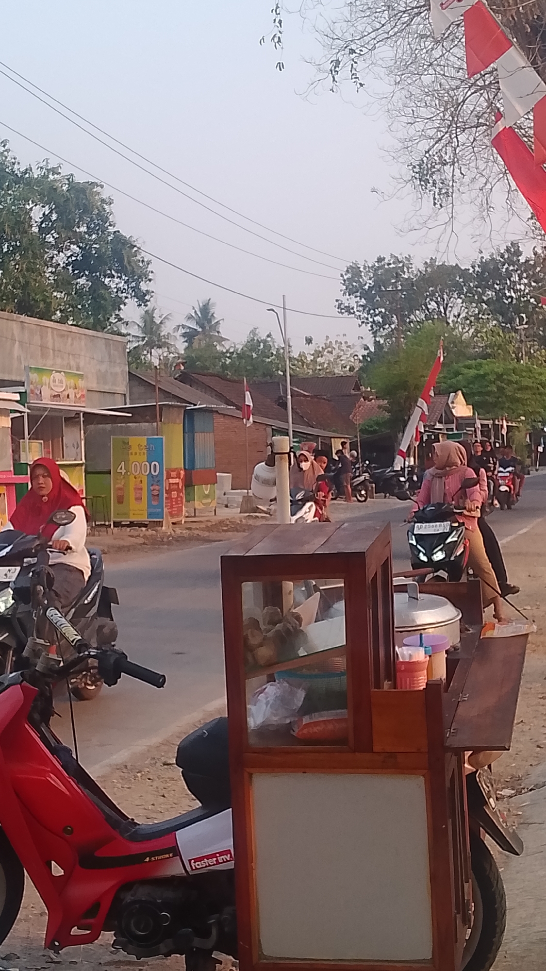 The image depicts a street scene in Karanglangu, Indonesia, during daylight hours, possibly late afternoon due to the warm lighting. The foreground features a food cart, likely selling snacks or drinks, mounted on a motorcycle. The cart has a wooden frame with glass display cases containing food items and a large metal pot. A red motorcycle with white and black accents is parked to the left of the cart, with a sticker reading "faster inv." visible on its side.
In the middle ground, a paved road runs horizontally across the image. Several people on motorcycles are present, suggesting typical street activity. On the left side of the road, there's a small building or shop with advertisements displayed, including one with the number "4.000" and images of drinks. A woman wearing a red hijab is riding a motorcycle.
Further back, a row of buildings lines the other side of the road, appearing to be residential or small commercial establishments. Indonesian flags, red and white, are visible, some mounted on poles and others attached to buildings or vehicles, indicating a possible national celebration or event. Palm trees and other lush green vegetation are scattered throughout the background, typical of the Indonesian landscape. The sky is clear with a pale, hazy appearance. The overall impression is of a bustling yet relaxed street environment in a semi-urban or suburban area of Karanglangu.