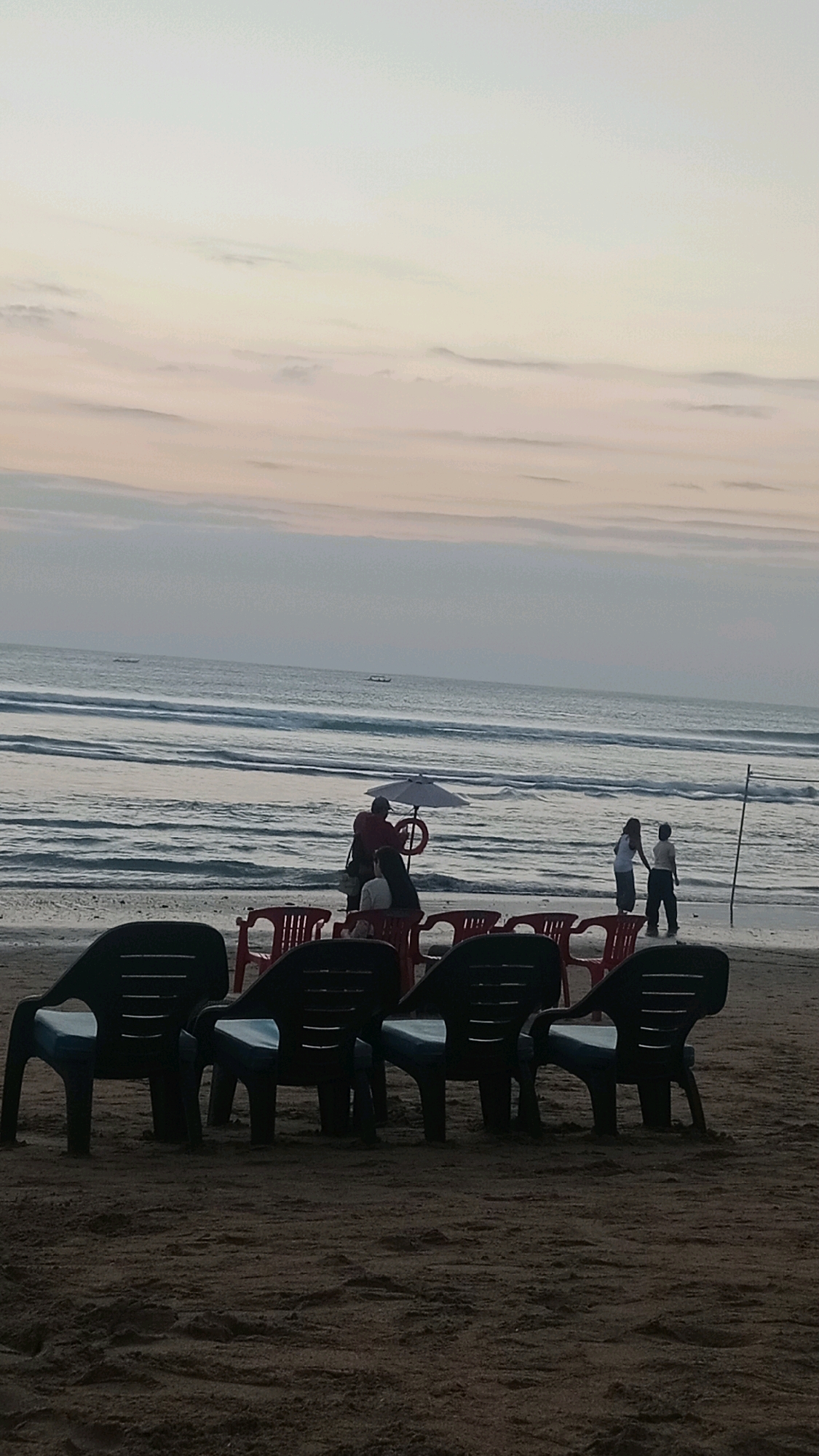 The image depicts a serene beach scene in Kuta, Indonesia, at dusk. The sky is a soft gradient of pale yellow and pink hues, with subtle cloud formations. The ocean stretches out to the horizon, with gentle waves lapping against the shore. In the foreground, several dark plastic beach chairs with blue seats are arranged on the sandy beach, some facing the ocean. Behind these chairs, a row of red plastic chairs is visible.
Further back, near the water's edge, a group of people is gathered. One individual, wearing a dark top, stands next to another person seated on a chair, holding a white umbrella. To their right, two other individuals, one in a white shirt and the other in a dark shirt, are standing. A thin pole with a horizontal bar is planted in the sand to the right of these figures. Two small boats are visible in the distance on the ocean. The overall atmosphere is calm and peaceful, capturing the tranquil beauty of Kuta beach as daylight fades.