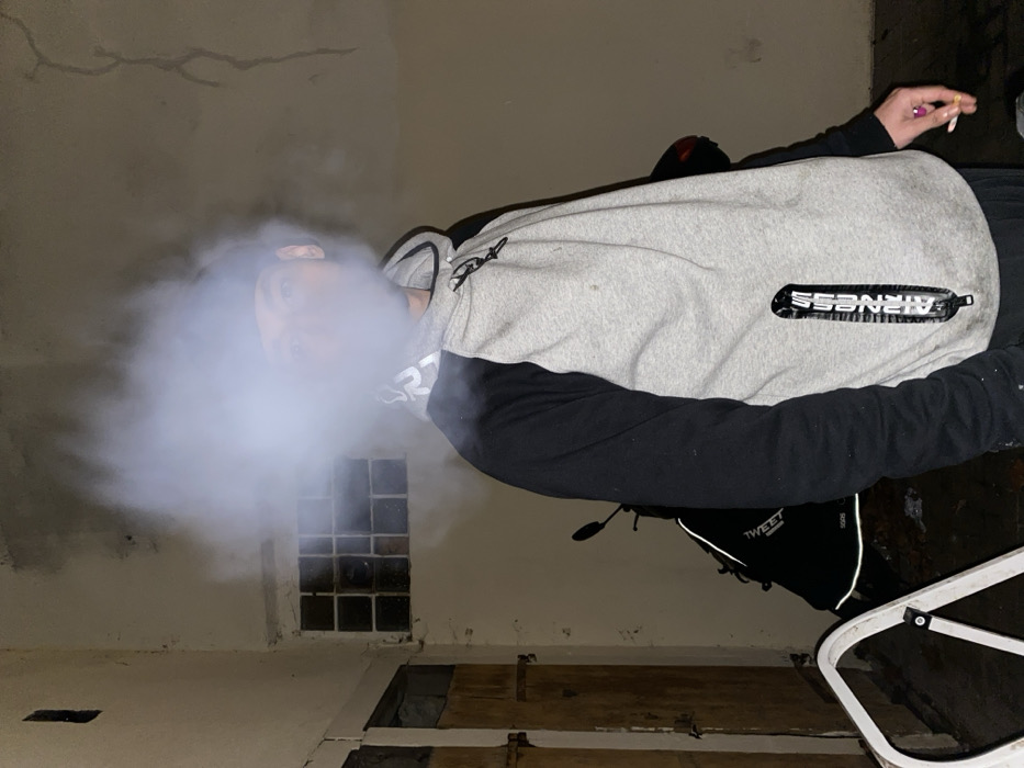 A person exhales a large cloud of vapor or smoke, obscuring their face. They wear a gray and black hooded sweatshirt with "AIRNESS" visible. The setting appears to be an old, unfinished room with damaged walls and floors. A partially visible window with broken panes is behind the person. A cigarette is held in the other hand.