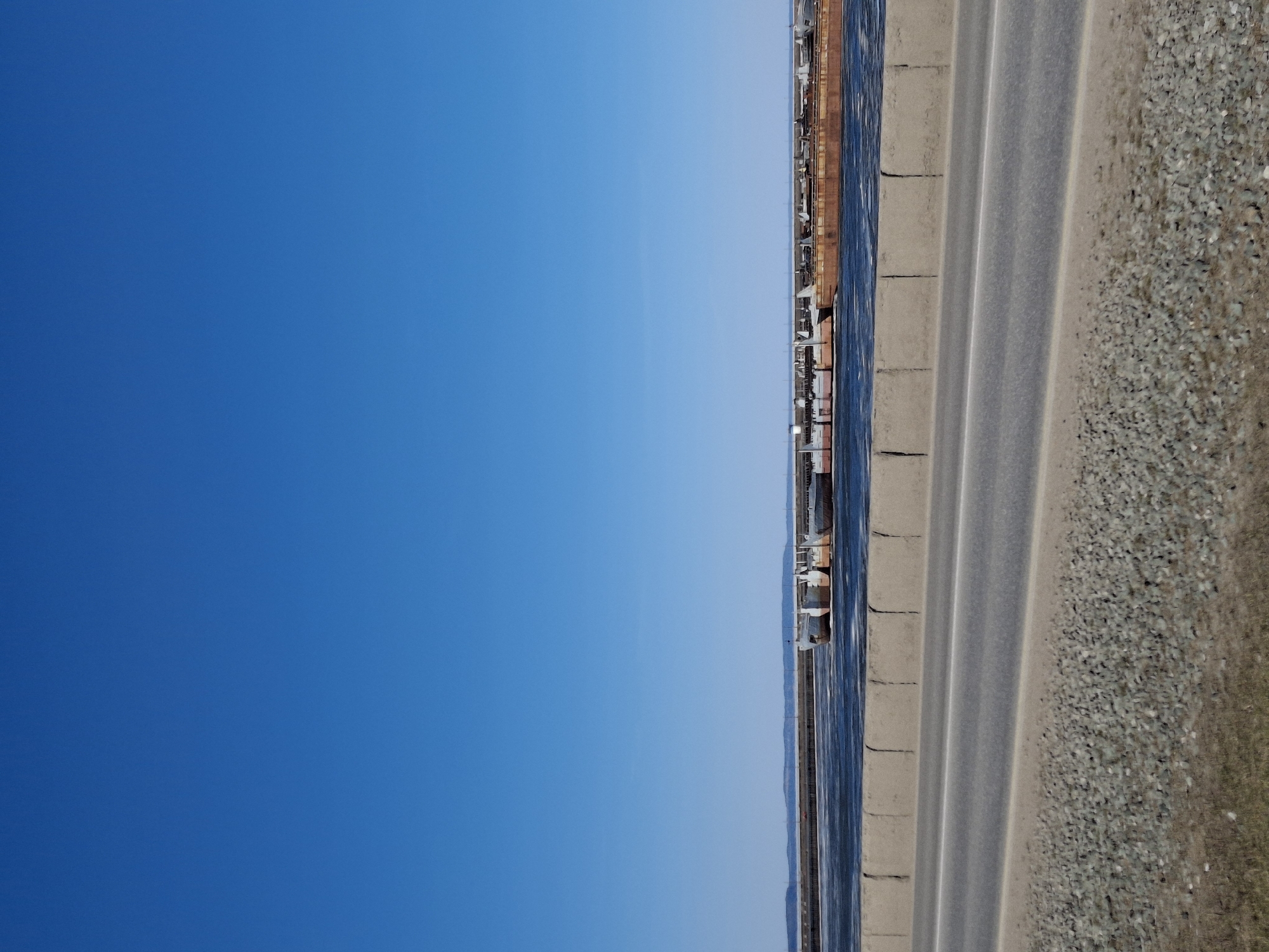The image shows a roadside scene in what appears to be Chaikovsky, Russia. A clear, blue sky dominates the left portion of the image. The right side features a stretch of asphalt roadway with multiple lanes and a shoulder comprised of gravel. Adjacent to the road is a concrete barrier, and beyond that, a body of water. In the distance, one can discern industrial structures, likely associated with a manufacturing or processing facility. The scene suggests a setting near an industrial area on the outskirts of a city.