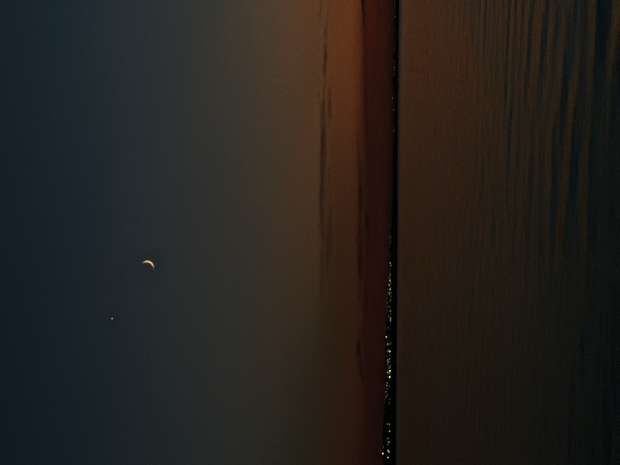 The image depicts a twilight scene over a body of water, likely a lake or river, with a distant city skyline illuminated by lights. The sky transitions from a deep blue at the top to shades of orange and red near the horizon, indicating sunset or sunrise. A crescent moon and a bright star or planet are visible in the upper portion of the sky. The water's surface is calm, reflecting the ambient light and the distant cityscape. The location is identified as the city of Kokshetau, Kazakhstan, suggesting this is a view of the city's waterfront or a nearby natural body of water. There are no people or specific actions visible in the image. The scene is primarily focused on the natural elements of the sky and water, with the city lights serving as a backdrop.