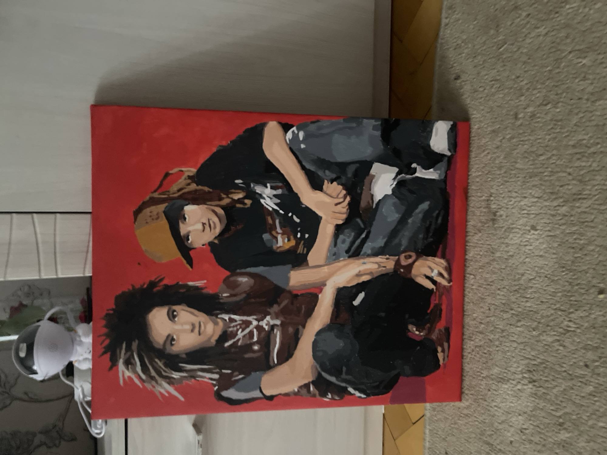 The image shows a painting of two individuals, both appearing to be male, crouching against a red background. The individual on the left has a distinctive hairstyle with spiky dreadlocks and is wearing a dark vest over a grey shirt and dark pants. The individual on the right wears a yellow baseball cap and a dark t-shirt with a graphic print, also in dark pants. Both subjects have their hands resting in their laps or on their legs. The painting is displayed on what appears to be a carpeted floor, with a wooden floor visible in the background. A white object with a spherical top and two wire-like appendages is partially visible in the upper left corner of the image, behind the painting. There is no discernible information in the image that indicates the location being in Марковский, Russia.