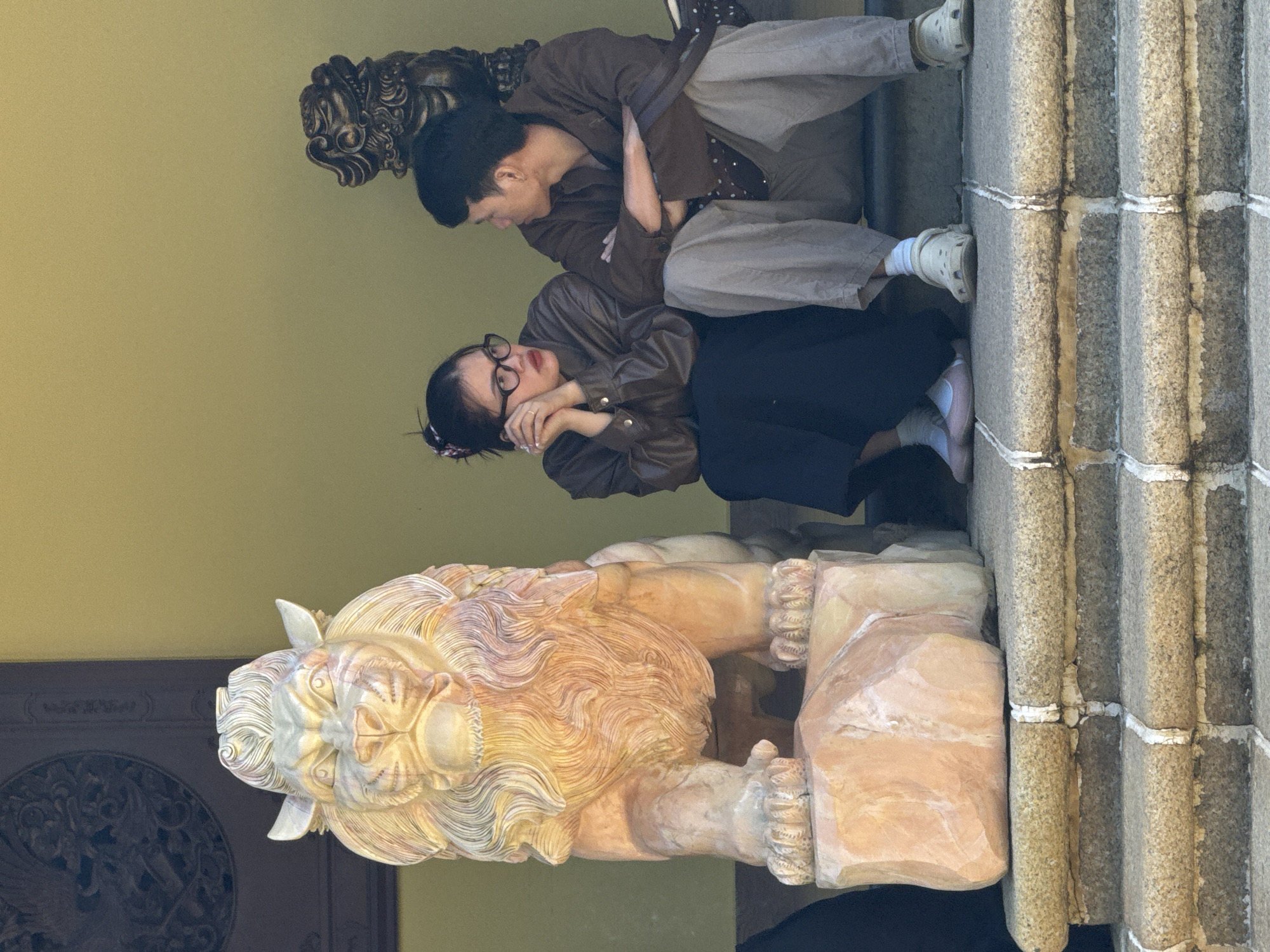 Two individuals, a male and a female, are seated on stone steps in front of a yellow building, likely a temple or a historic site, given the presence of a large, ornate stone lion statue to their left and a smaller dark statue of a similar creature behind the male. The female, wearing glasses and a brown jacket over a dark skirt, rests her chin on her hands. The male, in a brown jacket and grey pants, has his arms crossed. They appear to be in conversation. The lion statue is carved from a light-colored stone, possibly marble or sandstone, with intricate details in its mane and facial features. The steps are made of rough-hewn stone with white grout lines. The overall atmosphere suggests a peaceful and contemplative moment at a cultural landmark in Da Lat, Vietnam, known for its many temples and historical sites.