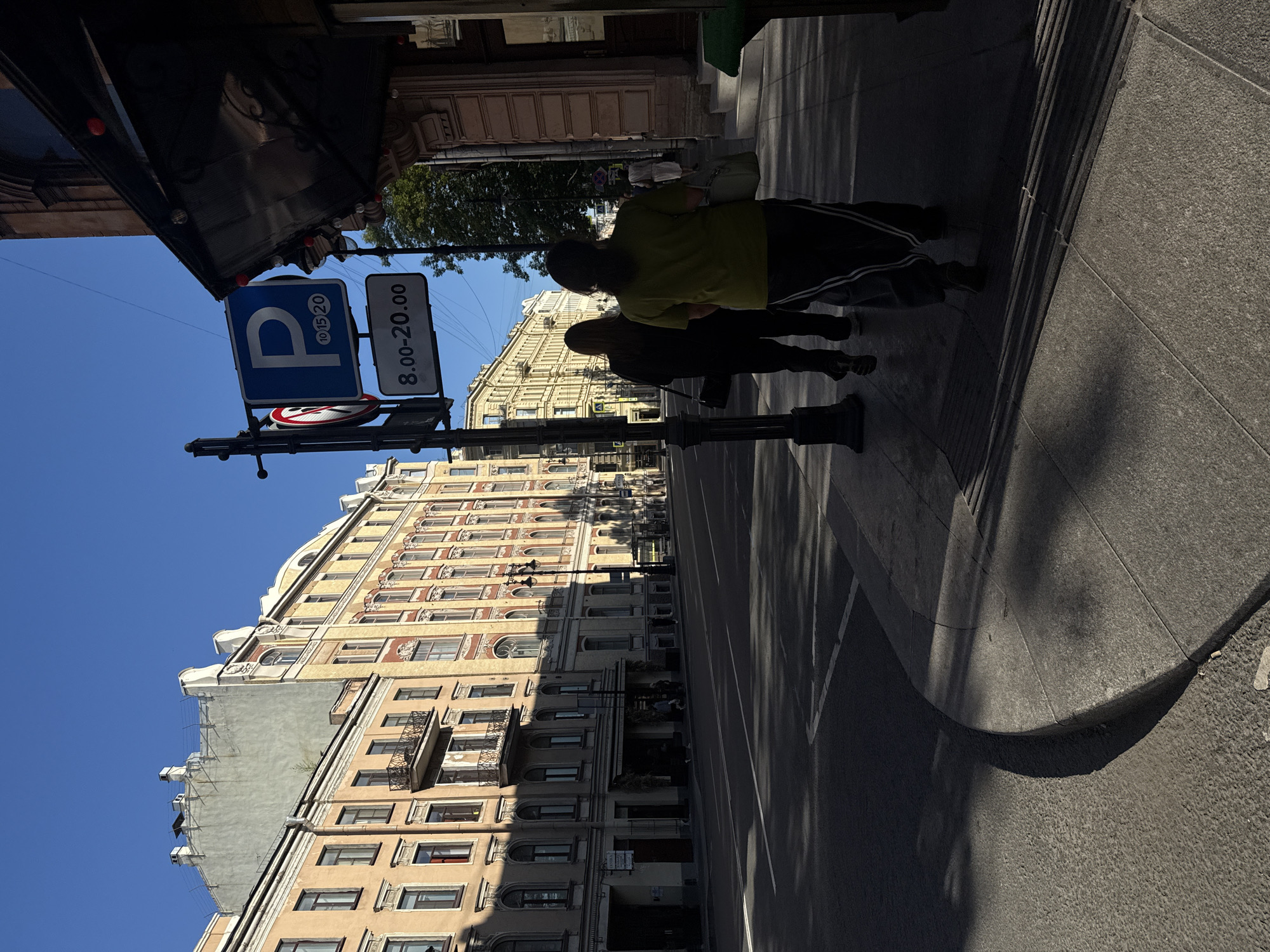 The image depicts a sunny street scene in Saint Petersburg, Russia, likely in the historic center, given the architecture. Two women are walking away from the camera on a sidewalk. The woman in the foreground wears a green t-shirt and dark pants with white stripes, and carries a light-colored bag. The woman behind her wears a black outfit and also carries a bag.
To their left is a street with visible lane markings. On the sidewalk, a lamppost supports a parking sign with a "P" and times "10 15 20", indicating parking regulations, and another sign below reads "8.00-20.00".
The buildings lining the street exhibit classic Saint Petersburg architecture, characterized by their ornate facades, tall windows, and pastel colors. The buildings are several stories high, with some featuring balconies and decorative cornices. The sky is a clear, bright blue, and the sun casts strong shadows across the street and sidewalk, suggesting it is either morning or late afternoon. The overall impression is of a quiet, orderly urban environment.
