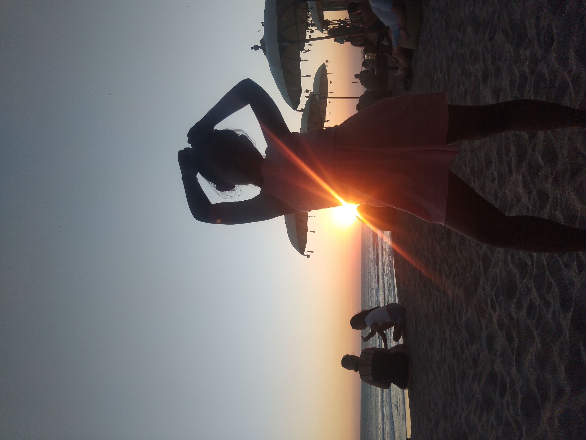 This image, captured in North Kuta, Indonesia, depicts a serene sunset scene on a beach. The sun, a bright, glowing orb, is positioned low on the horizon, creating a prominent lens flare that streaks across the frame. Its vibrant orange and yellow light illuminates the lower sky, gradually fading into a pale blue-grey at the top.
In the immediate foreground, the silhouette of a person, likely a woman or girl, stands facing the setting sun. She is wearing a dress and has her arms raised with hands behind her head, creating a dynamic pose against the bright light. Her dark form is sharply defined against the luminous backdrop.
Further back, near the water's edge on the right side of the frame, two other silhouetted figures are visible. One person is kneeling or bending down on the sand, while another sits or kneels slightly behind them. The gentle waves of the ocean can be seen lapping at the shore. In the background, several large beach umbrellas are scattered across the sand, suggesting a beach resort or a well-frequented area. The sand in the foreground shows subtle textures. The overall mood is peaceful and reflective, characteristic of dusk at a beach destination. No visible text is present in the image.