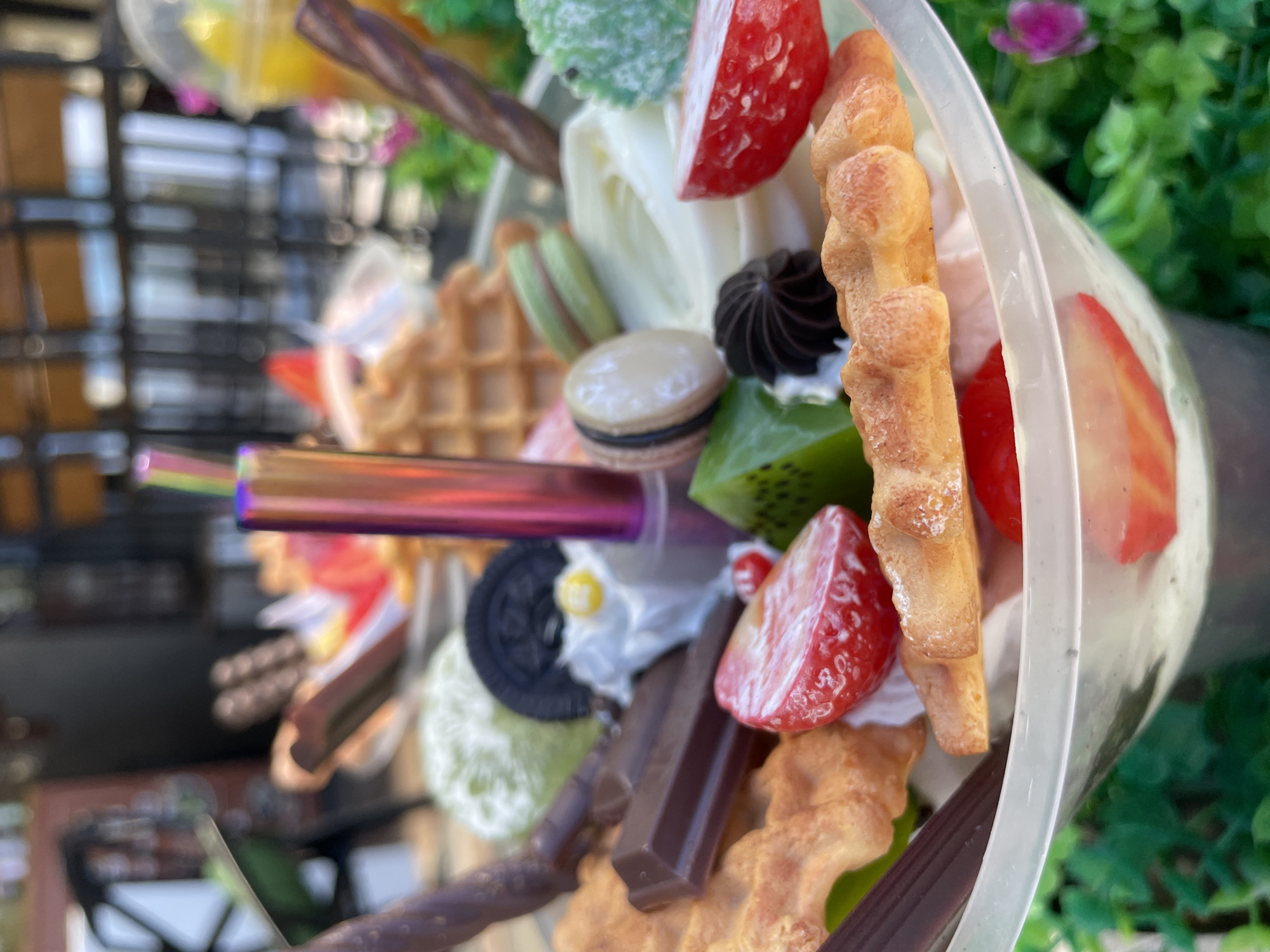 This is a close-up, eye-level shot of a dessert in a clear plastic cup. The dessert is elaborately decorated with a variety of items including waffles, chocolate bars, strawberries, macarons, whipped cream, and what appears to be ice cream. A colorful, iridescent straw stands vertically in the center of the cup. The background is blurred but shows what looks like an outdoor seating area with plants and possibly tables and chairs. The lighting suggests it is daytime. No people are visible in the frame. The image does not provide any information about the city of Sochi, Russia.