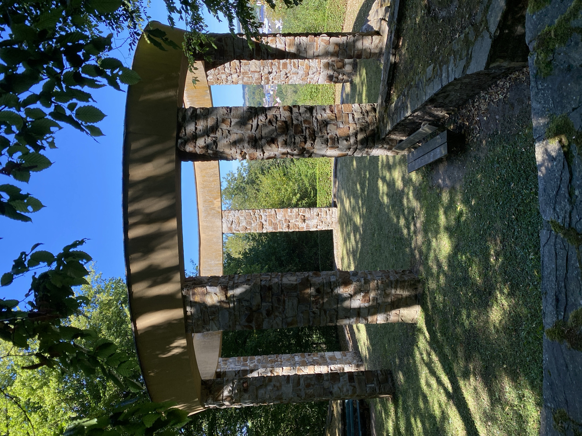 An open-air stone structure with a circular roof supported by stone pillars stands on a grassy knoll under a clear blue sky. The structure appears to be a pavilion or monument, with stone benches lining its perimeter. Trees and foliage surround the area, and in the distance, a town with buildings can be seen nestled in a valley. The scene is lit by sunlight, casting shadows across the grass and the stone elements of the structure. The location is the Johanniskreuz lookout tower, a landmark in the city of Altenkirchen, Germany.
