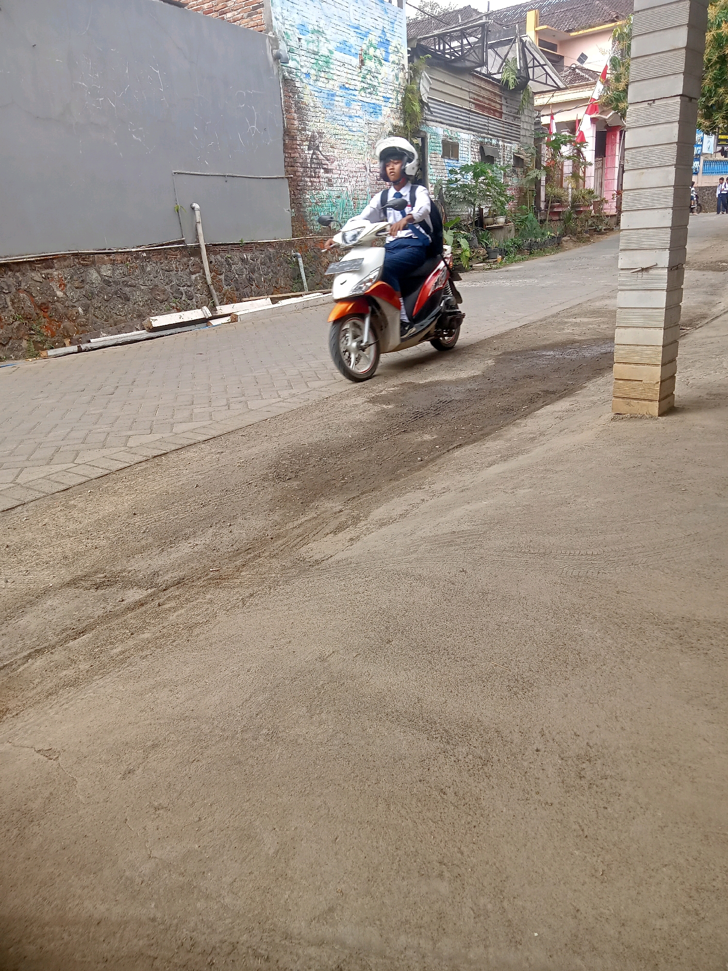 A person wearing a white helmet and a school uniform is riding a white and orange scooter on a street in Pule, Indonesia. The scooter is moving from left to right in the frame. The street is paved with interlocking bricks on the left side and asphalt on the right. There are buildings and a brick wall with a mural on the left side of the street. On the right side, there is a tall, square pillar made of stacked concrete blocks and some trees. In the background, there are more buildings and what appears to be a residential area. The lighting suggests it is daytime, possibly overcast.