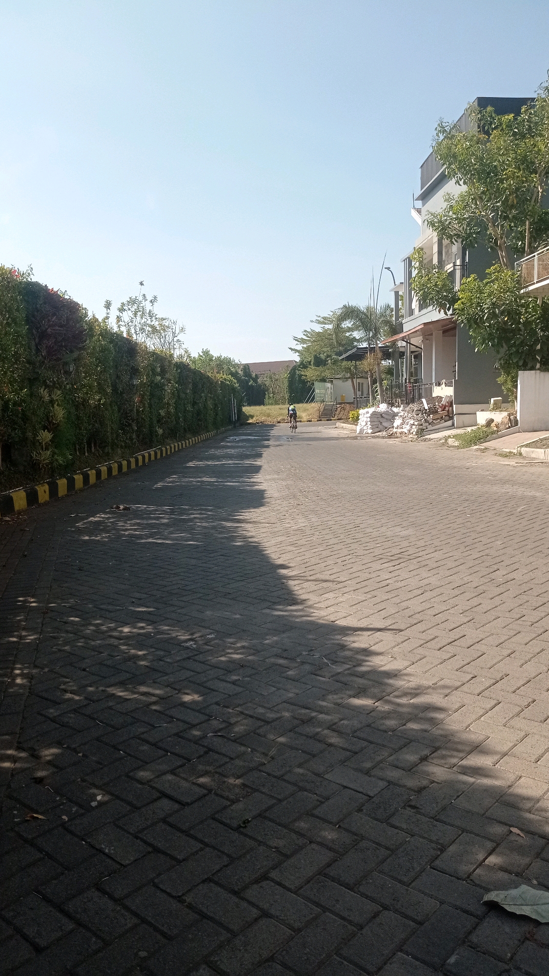 A lone cyclist rides a bicycle on a paved road in a residential area, possibly in the city of Batu, Indonesia. The road is made of dark grey interlocking bricks and is lined on the left by a tall, dense green hedge with yellow and black striped curbing. On the right side of the road, there is a modern-looking building with multiple stories, balconies, and trees. The building appears to be gray with white trim and a dark roof. In front of the building, there are piles of sandbags and construction materials, suggesting ongoing development or renovation. The sky is a clear, pale blue with no clouds. Sunlight casts dappled shadows of trees across the road. The overall scene suggests a peaceful, sunny day in a suburban or developing urban environment.
