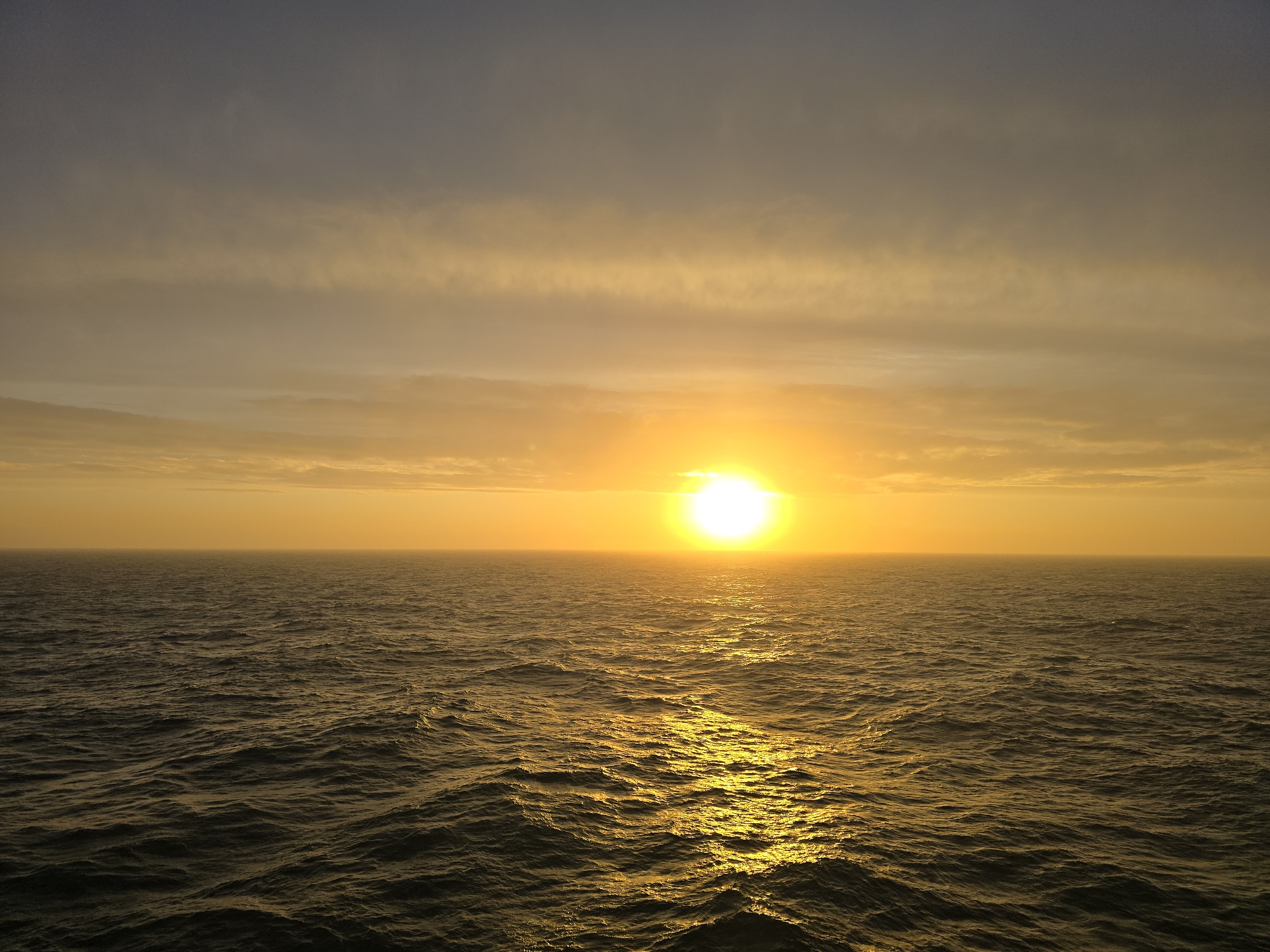 The image shows a sunset over the ocean. The sun is positioned in the center of the horizon, radiating a bright yellow and orange light. The sky above the sun is filled with horizontal layers of clouds, transitioning from darker shades at the top to lighter, golden hues closer to the horizon. The ocean surface is rippled and reflects the golden light of the setting sun.
The location of the scene is unknown.