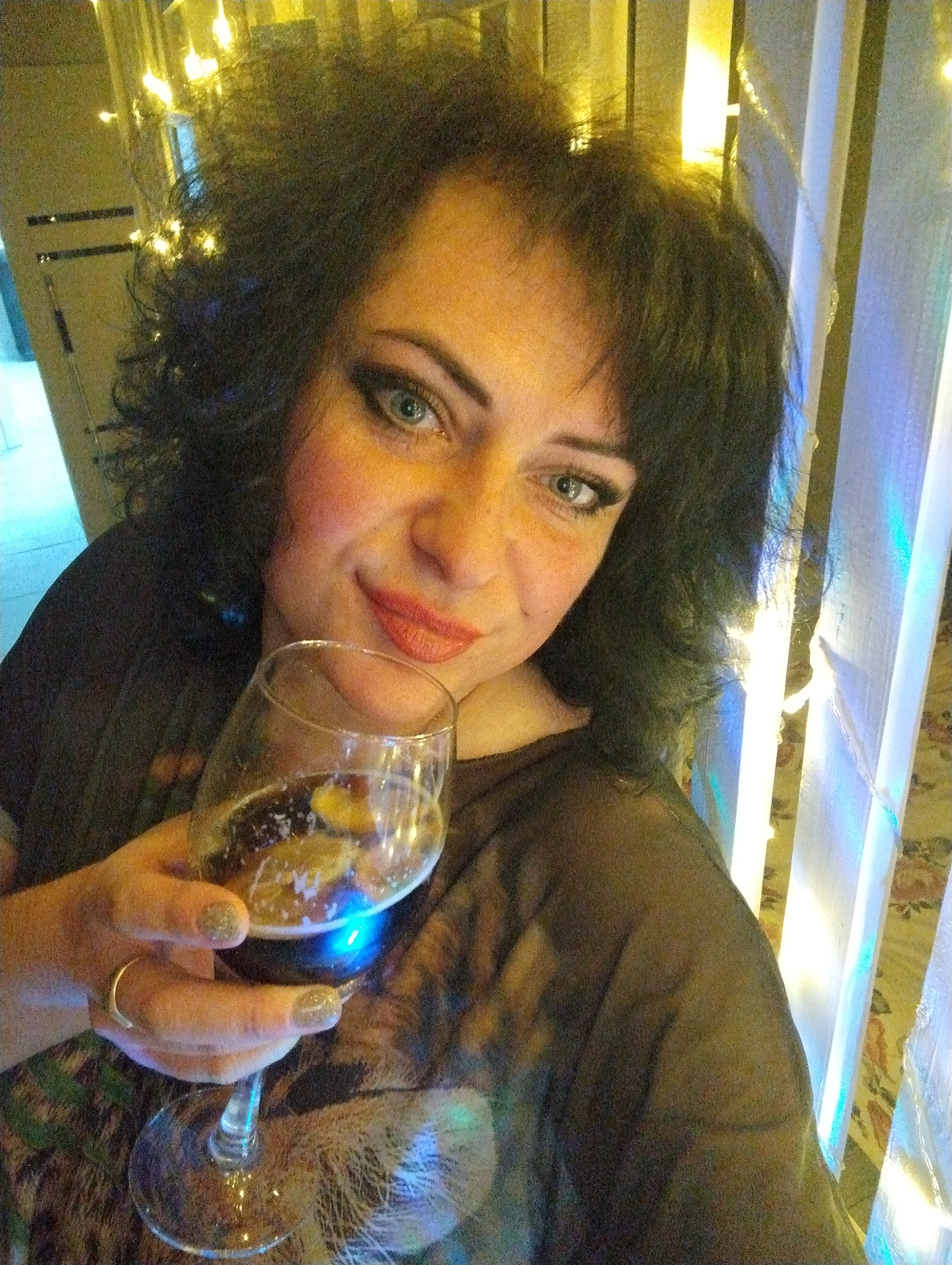 A woman is holding a wine glass filled with liquid and lemon slices. She has dark, curly hair, light eyes, and is wearing lipstick. She is wearing a ring on her finger. There are illuminated decorations in the background, including hanging lights and lighted bars. The exact location within Grishanki, Russia, cannot be determined from the image.