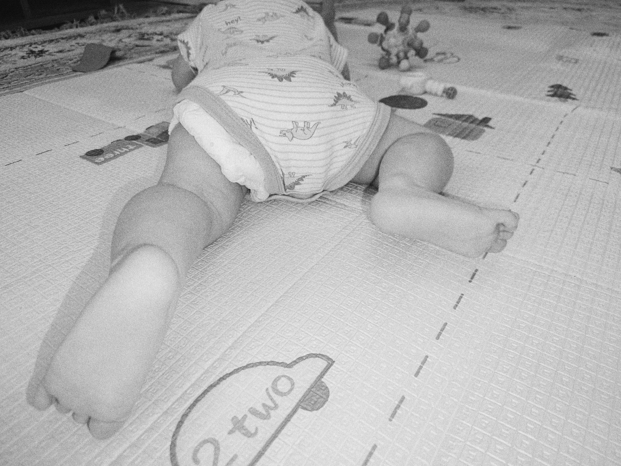 The image shows the lower body of a baby crawling on a play mat. The baby is wearing a diaper and a onesie with a dinosaur pattern and the word "hey!". The play mat has a textured surface and printed markings, including dashed lines and the word "two" within a speech bubble shape. In the background, there is a fuzzy toy and a patterned rug. The image is black and white and appears to be a candid shot. There is no information within the image or provided context that would indicate the location as Tobolsk, Russia.