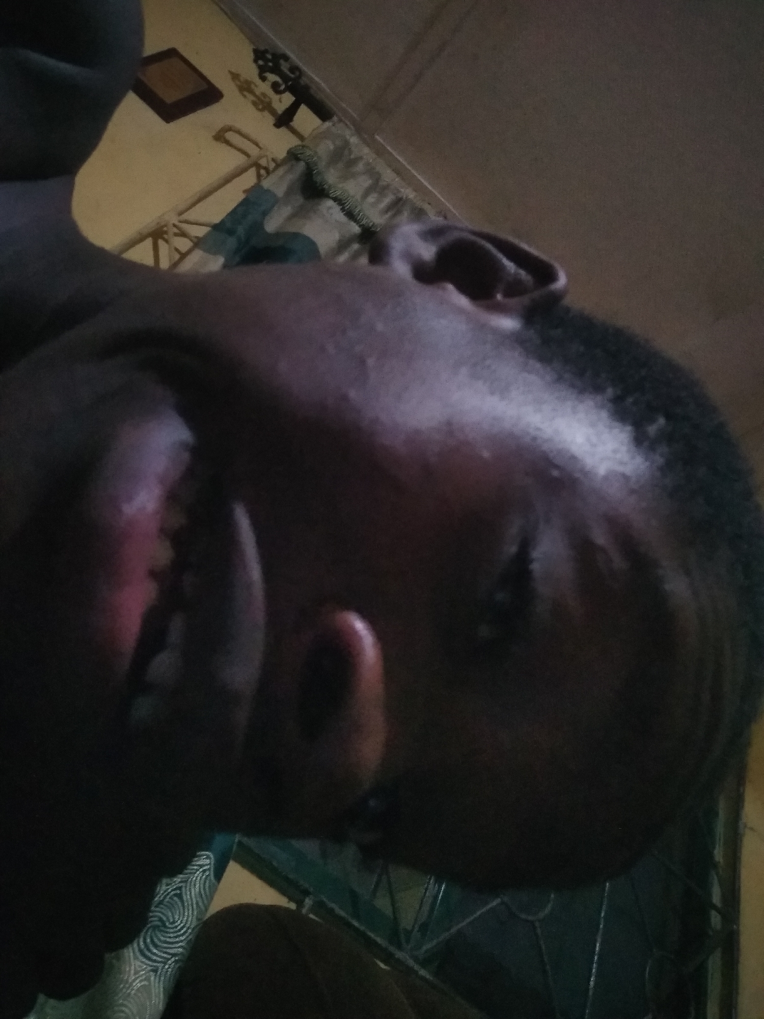 A low-angle, off-center photograph depicts a person's face, with the head tilted back. The individual has dark skin and short, dark hair. Facial features such as the ear, nose, and teeth are visible, although partially obscured by the angle and lighting. The background shows elements of a room, including a window with a curtain, part of a ceiling with a small square object (possibly a picture frame), and a decorative metal object. The location is not specified within the image.