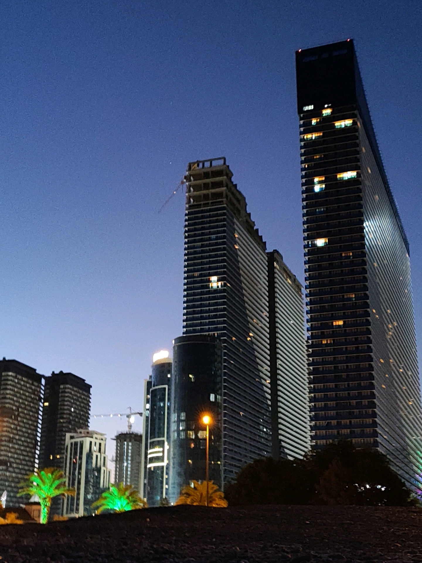 The image depicts a modern cityscape at dusk, likely in Batumi, Georgia, given the architectural style and palm trees. Dominating the scene are several tall, contemporary buildings. On the right, a very tall, sleek skyscraper with a dark top and illuminated windows stands prominently. To its left, another skyscraper of similar height features a textured facade with many windows, some lit. Further to the left, a building under construction is visible, with exposed framework and a crane extending upwards. In the foreground, a dark, textured ground surface, possibly a path or embankment, leads the eye towards the buildings. Lush palm trees, one of which is vibrantly illuminated with green light, punctuate the middle ground, adding a tropical element to the urban environment. The sky above is a deep, clear blue, transitioning towards darkness as night approaches. A streetlamp with a warm orange glow is visible in the middle right of the frame. No people are discernible in the image.