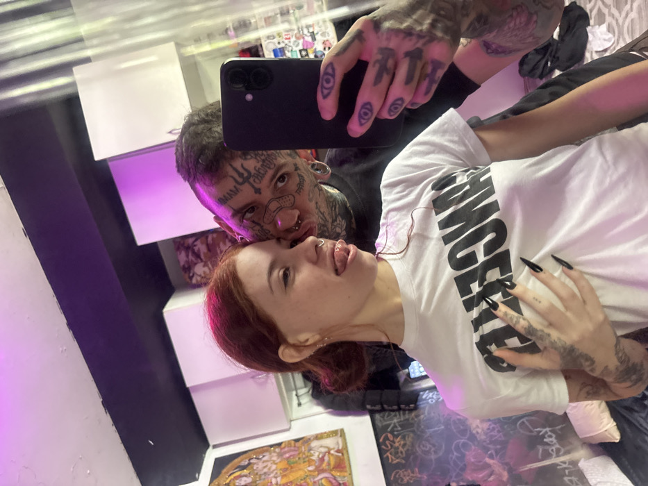 The scene takes place indoors, within a private residence in Buenos Aires, Argentina. No specific landmarks or street-level details are visible to further pinpoint the exact location within the city.
The image is a mirror selfie featuring two individuals, a female and a male, bathed in purple ambient lighting. The female, positioned on the left, has reddish-brown hair styled into two buns and multiple facial piercings, including a septum ring and earrings. She wears a white short-sleeved t-shirt with the black word "CANCER" printed horizontally across the chest. Her left hand, adorned with long, pointed black fingernails and various tattoos, rests on her midsection. She looks towards the camera with her tongue extended.
The male, positioned behind and to the right of the female, has numerous dark tattoos covering his face, neck, and arms. His facial tattoos include symbols on his forehead, a cross-shaped element, a bandage icon on his cheek, and visible text elements such as "MAM" and "calita." He has multiple facial piercings, including lip and nostril piercings, and gauges in his earlobes. His dark hair is styled in small dreadlocks or twists. He holds a black smartphone in his right hand, which is also heavily tattooed with "eye" motifs on his fingers, using it to capture the mirror image. He looks directly into the phone's reflection.
The background includes a framed depiction of a multi-limbed deity, possibly Hindu, in golden/orange hues, displayed on the left wall. Below this, a dark surface exhibits white graffiti-like markings. White overhead cabinets or shelves are visible in the upper-center background. Behind the male's head, a patterned pillow or fabric is visible. The reflective surface on the right, functioning as the mirror, shows reflections of small, indistinct items, possibly stickers. A bed or sofa with dark bedding and a white pillow is partially visible in the lower-left foreground.