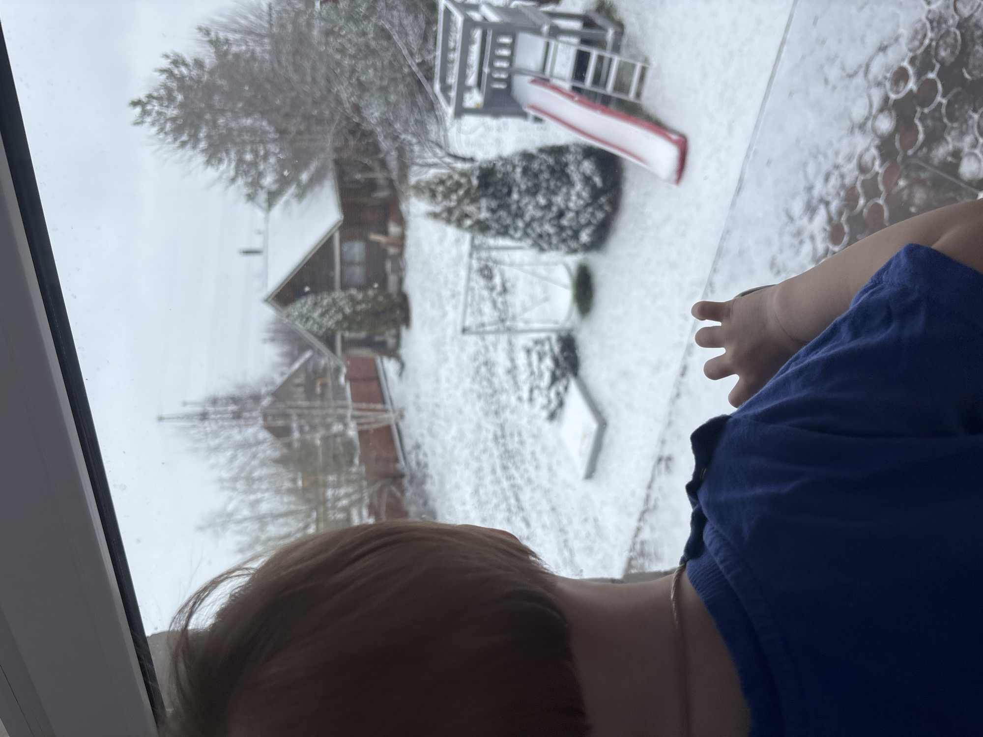 A child is observing a snow-covered landscape through a window. The scene outside includes houses, trees, and a play structure with a red slide, all covered in snow. The child is wearing a blue shirt and is pointing at the snow-covered landscape. The location is Ферма, Russia, as that was stated by the prompter.