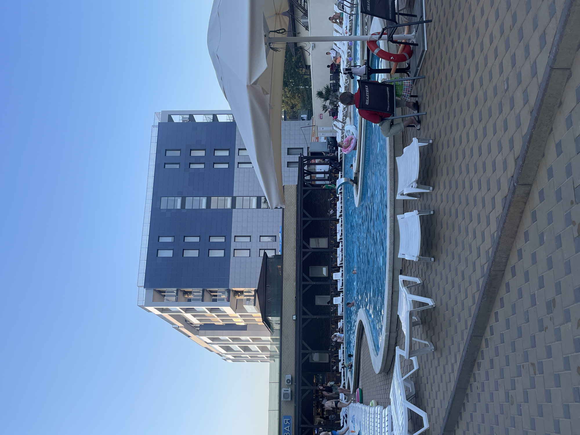 This media file depicts an outdoor swimming pool area at a resort in Anapa, Russia. The scene is bathed in the warm light of either late afternoon or early evening.
The main subjects are people relaxing and swimming. Several individuals are seated in lounge chairs on the paved patio surrounding the pool, while others are in the water. One person is visible in a red shirt sitting at a table with a black board, possibly engaged in work or leisure. A child can be seen in a pink inflatable ring in the pool. The pool itself is a vibrant blue, with curved edges and white coping.
In the background, a modern hotel building with a blue and gray facade dominates the view. The building features numerous rectangular windows. Adjacent to the hotel is a darker, multi-story structure with a dark, latticed facade, possibly housing amenities or rooms.
Notable details include the presence of white plastic lounge chairs, a red life preserver, and a large white umbrella providing shade. The paving stones on the patio are in a patterned arrangement of grey and blue-grey squares. The sky is a clear, pale blue, suggesting fair weather.
The overall atmosphere appears relaxed and leisurely, indicative of a vacation or resort setting. The visible text "Балтика" (Baltika) is present on the black board held by the person in the red shirt, which is a Russian beer brand. The word "BAR" is also visible on a sign on the darker building.