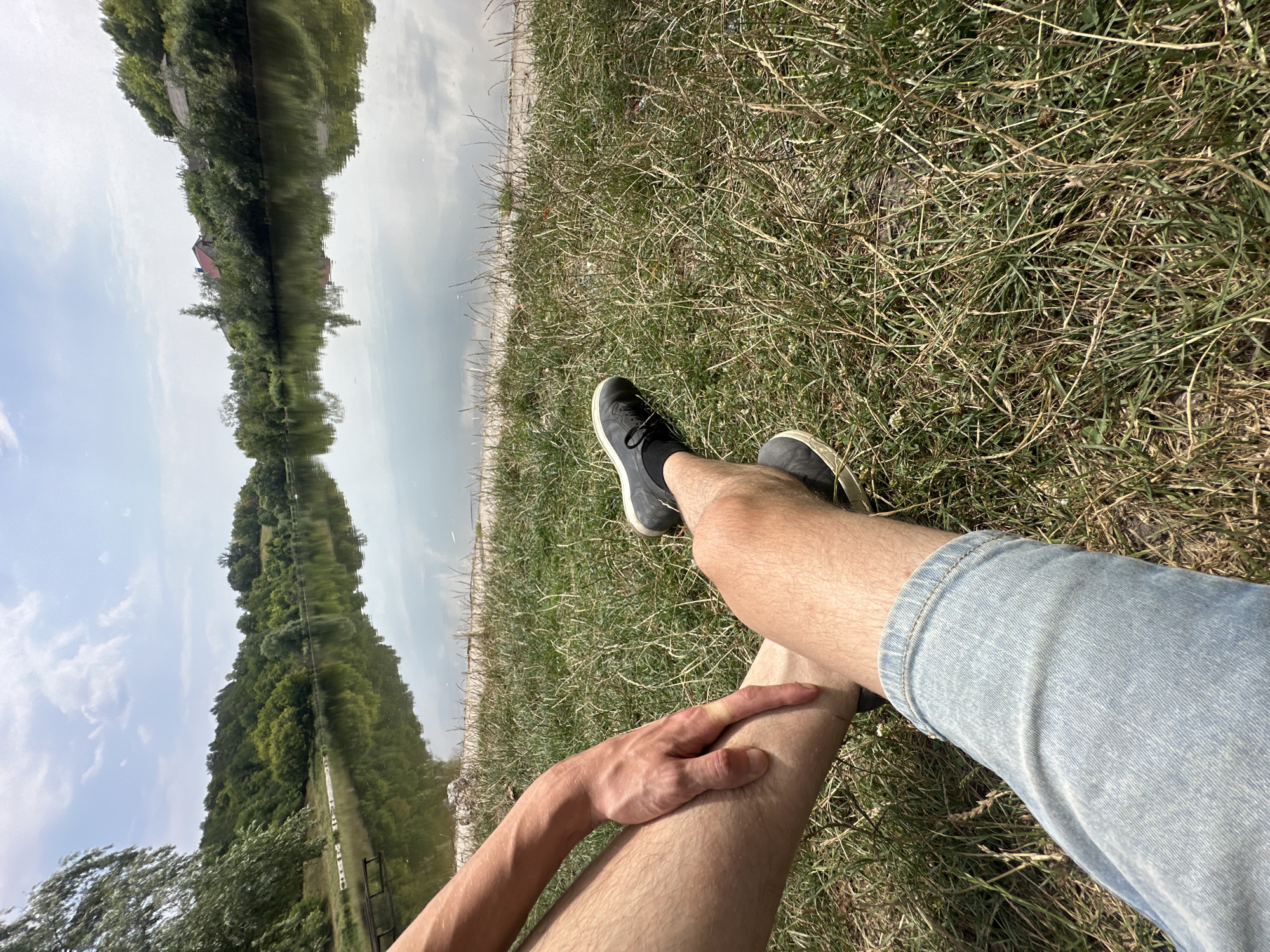 The image depicts a low-angle, first-person perspective of two pairs of legs and one hand resting on the grass by the side of a calm lake. The legs are positioned in the foreground, with one pair clad in light blue denim shorts and the other in dark athletic shoes and black socks. The hand, resting on the knee of the individual in shorts, appears to belong to the person wearing the shoes. The background features a tranquil lake with a reflection of a tree-lined shore and a partly cloudy sky. Scattered on the shoreline are small rocks and some sparse vegetation. Further back, the shore is densely populated with trees, and a few houses with red roofs are visible among the foliage. A wooden structure, possibly a pier or seating area, is partially submerged in the water on the left side of the frame. The overall environment suggests a rural or semi-rural setting, likely on the outskirts of Makiv, Ukraine, given the characteristic vegetation and the presence of houses.