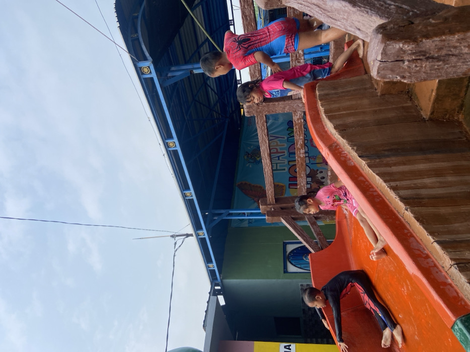 The image shows four children playing on a water slide at a waterpark in Kecamatan Tanggulangin, Indonesia, located at coordinates -7.497 latitude and 112.684 longitude. One child is sitting on the slide, another child is climbing the steps to the slide, and two children are standing by the slide. The slide is orange and red and is made of plastic. The steps are made of wood. There is a wooden fence behind the slide with a sign that says "Happy Holidays." The children are wearing swimwear. The sky is cloudy and the weather is overcast. The ground is wet.