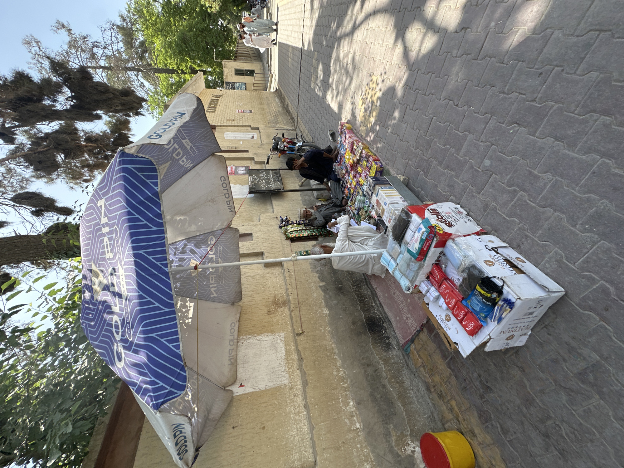 A medium shot, taken at eye level, shows a street vendor in Quetta City Tehsil, Pakistan, displaying goods on a sidewalk next to a building. The vendor is positioned behind a table laden with an assortment of items, including snacks, candy, and face masks, all neatly arranged for sale. An umbrella, branded with "GOLDPIN", provides shade over the stall.
Two people are visible, seemingly involved in the vending activity. They appear to be tending to the stall, possibly serving customers.
The sidewalk is paved with interlocking blocks, and the building beside the vendor is an old style construction with plaster walls. A motorcycle is parked behind the vendor. The overall environment suggests a street scene.