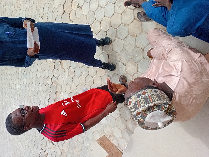 The scene, located in Kwazailewa, Nigeria, depicts an outdoor gathering of at least four individuals on a paved surface composed of light-colored hexagonal pavers and a lighter, possibly tiled, area in the lower left. The lighting indicates daylight.
In the foreground, an older male is seated directly on the paved ground, wearing a light peach-colored patterned long-sleeved garment and a multi-colored patterned cap with a white central dome. He gestures with his raised right hand, palm open, and looks upwards towards the standing individuals. To his right, partially visible, is another seated individual wearing blue trousers and dark slip-on shoes.
Standing to the left, a male in dark-framed glasses and a red Adidas t-shirt with black shoulder stripes is smiling broadly and looking towards the seated older male. He wears brown sandals.
To the right, another individual stands, wearing a dark blue traditional long garment over dark trousers and black athletic shoes. This individual holds a folded white document in both hands at waist level; a blue pen is visible in their chest pocket. The head of the individual in the dark blue garment is not visible.
The interaction suggests an ongoing discussion or address among the group.