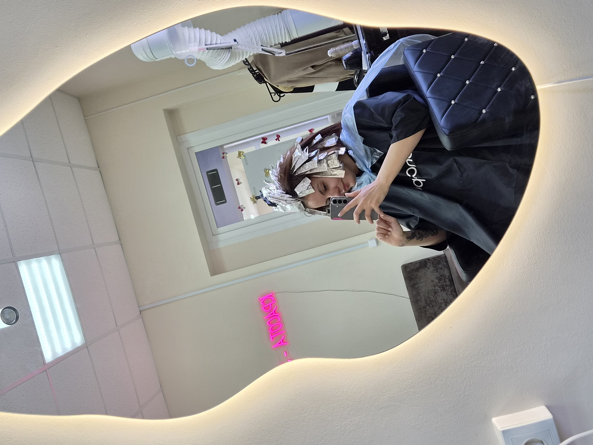 A mirror reflects a woman in a salon setting, likely in Chaikovsky, Russia. The woman is seated, undergoing a hair coloring process involving foil application. She holds a smartphone, presumably taking a selfie. A dark-colored salon cape covers her clothing. In the background, a salon chair with a quilted pattern is visible. A pink neon sign reading "COSMOCALY" is displayed on the wall, alongside a ceiling with fluorescent lighting and an air vent. A beige bag hangs on a chair or equipment behind the woman.