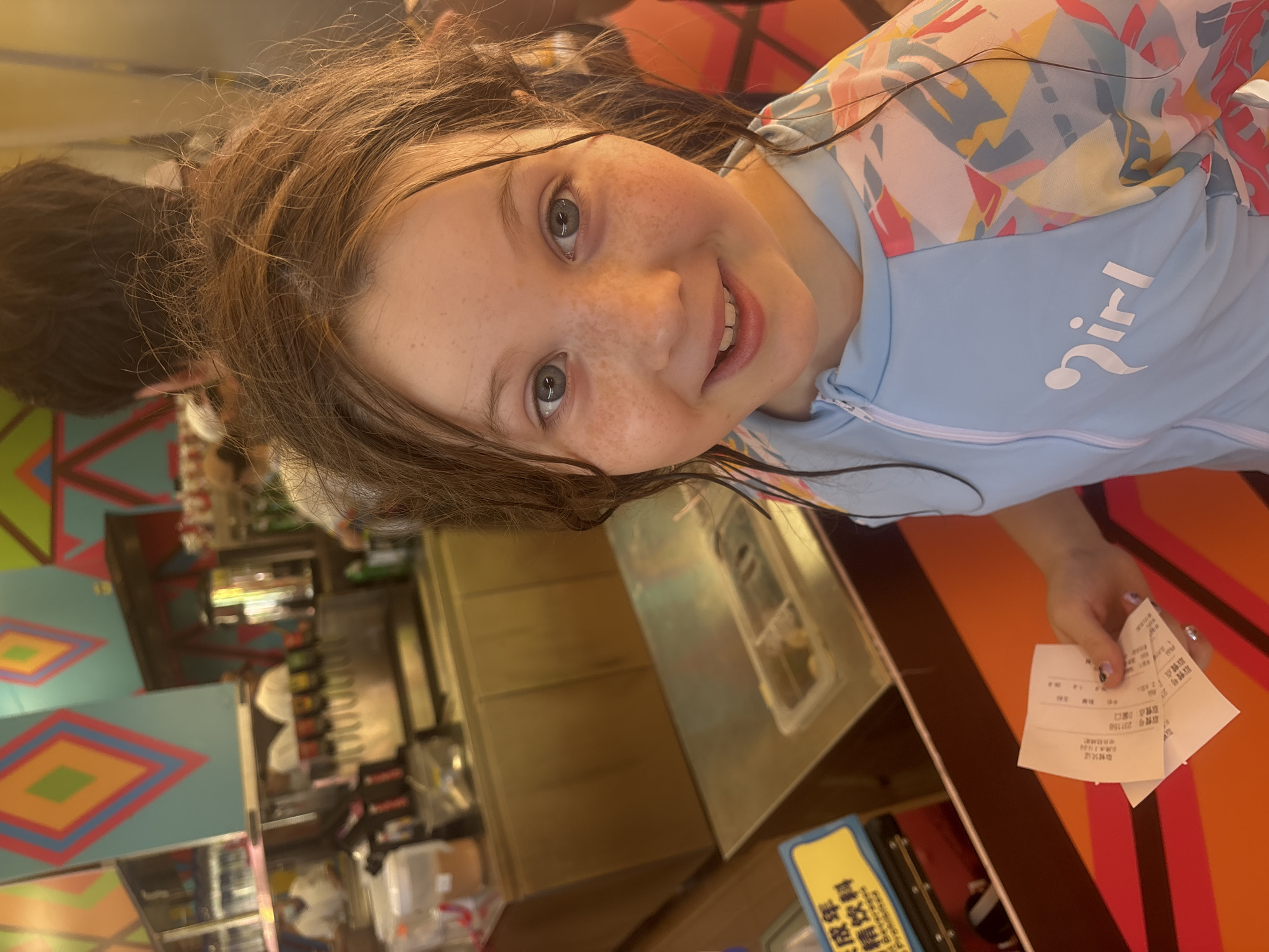 A young Caucasian girl with freckles and wet brown hair is looking at the camera and smiling. She is wearing a light blue rash guard with a colorful abstract pattern on the sleeves and the text "Girl" printed on the front. She is holding a receipt in her right hand. In the background, there is a food service counter with drink dispensers and cabinetry. A yellow sign with Chinese characters is visible to the left. The walls are decorated with colorful geometric patterns. The overall environment suggests a casual dining or fast-food establishment, potentially in the Panyu District of China, given the presence of Chinese text on signage and receipts.