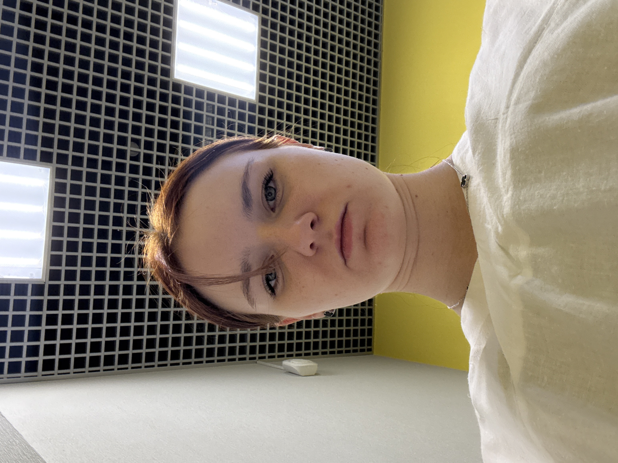 The image shows a person with fair skin and short reddish-brown hair. The person has light blue eyes and appears to be looking directly at the camera. They are wearing a light-colored, possibly white, garment. The background consists of a black and white checkered tiled wall with two illuminated square panels, and a yellow-painted wall. The image appears to have been taken in an indoor setting. A white object, potentially a mouse, rests on a gray surface. The location information provided indicates a city and state of "Unknown, Unknown."