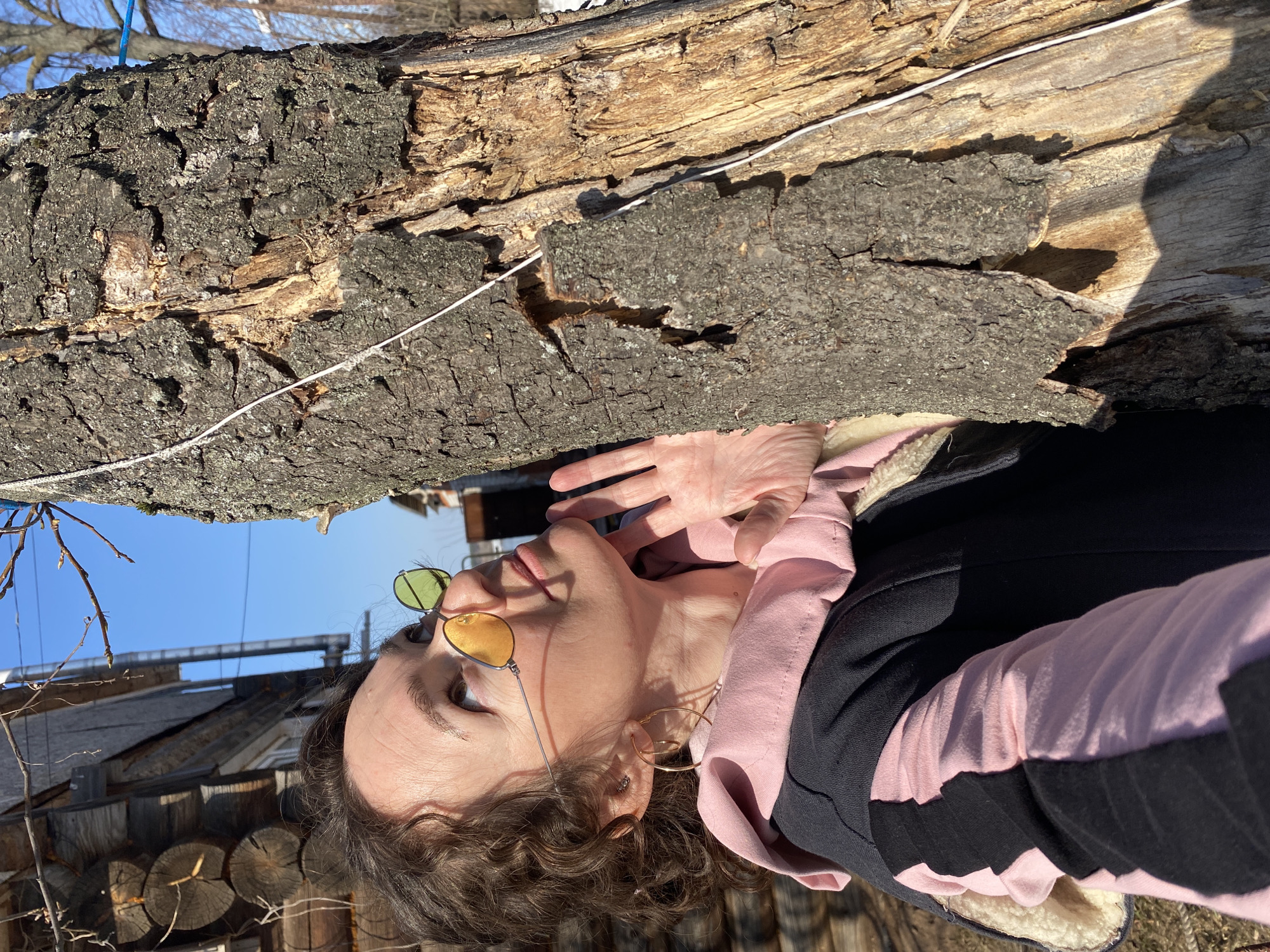 The image shows a woman wearing sunglasses and a pink and black jacket standing outside in Ольховка, Russia. A large log lies across the top of the frame, showing its bark texture. A thin white cord is attached to the log. In the background, partially obscured by the woman and the log, a wooden structure (possibly a house or shed) is visible under a bright blue sky. The woman's hand is raised near her face. The orientation of the photo is rotated, making the sky appear to be below the log. The environment seems to be a sunny outdoor setting in a rural or semi-rural location.