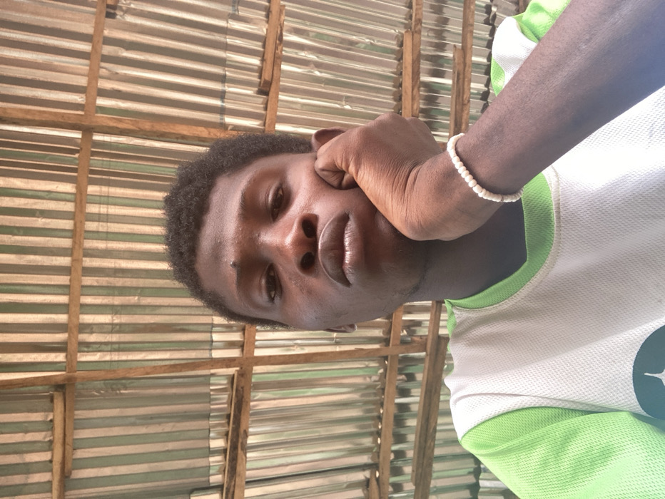 A dark-skinned male, located in Agege, Nigeria, is shown from the chest up, looking directly at the camera. He has short, dark, curly hair and a small mole superior to his right eye. His right hand is positioned with the palm against his chin. A white beaded bracelet is present on his left wrist. He is wearing a white t-shirt with light green trim on the collar and shoulders. The background consists of corrugated metal sheets, likely a roof or wall, supported by horizontal and vertical wooden beams.
