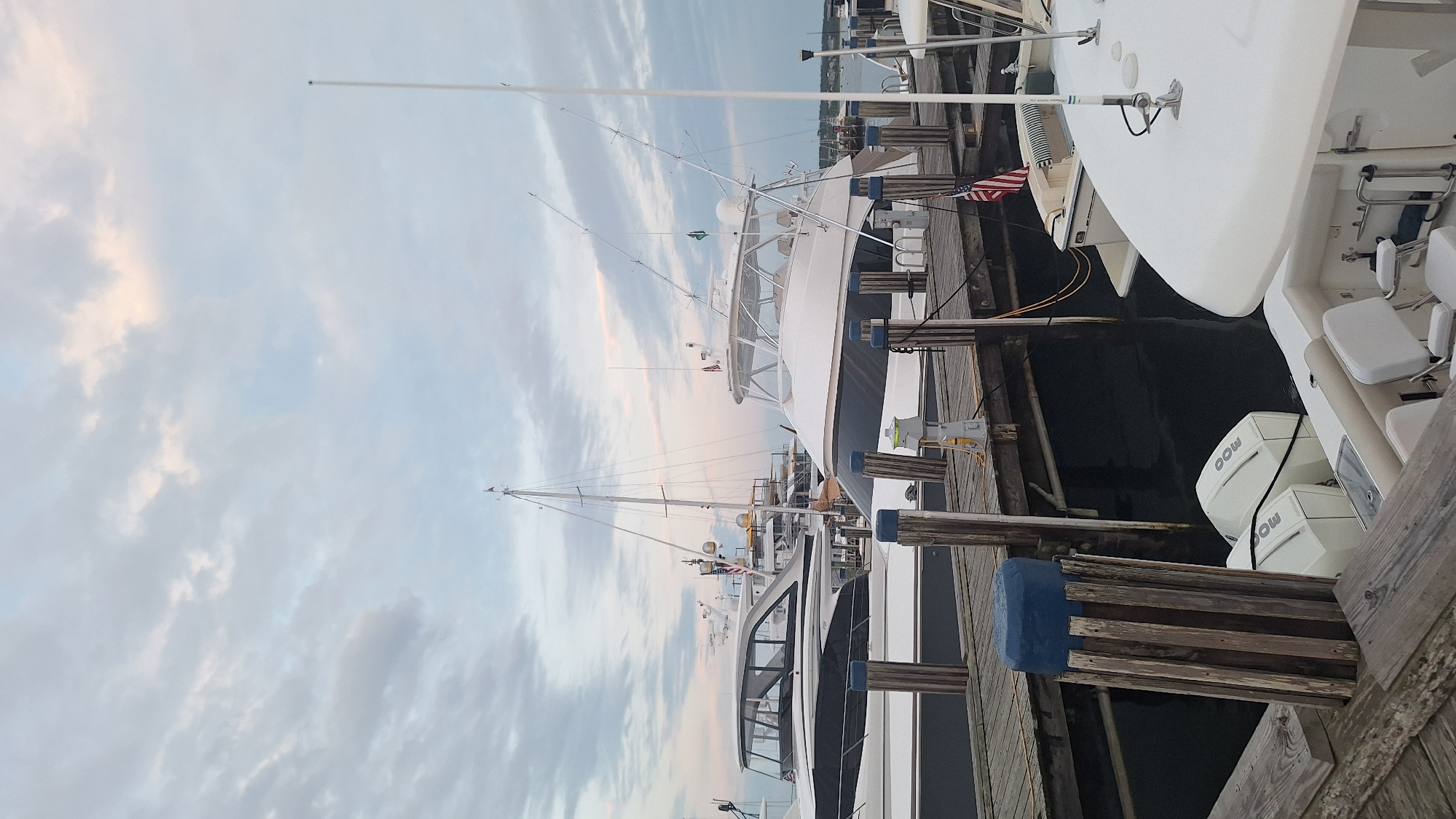 The image depicts a marina in Nantucket, United States of America, with multiple yachts docked. In the foreground, a wooden dock runs horizontally, with several pilings, one capped with a blue object, supporting it. Two outboard motors, branded "Mercury," are visible attached to the stern of a white boat. Beyond the foreground, another wooden dock is present, with a white yacht docked to it, featuring fishing rods extending from its superstructure. To the left, a larger white yacht with a dark hull and enclosed cabin is docked. In the background, more yachts and a sailboat are visible. The sky is overcast with soft clouds, suggesting either early morning or late evening. The overall scene is one of a tranquil marina setting.