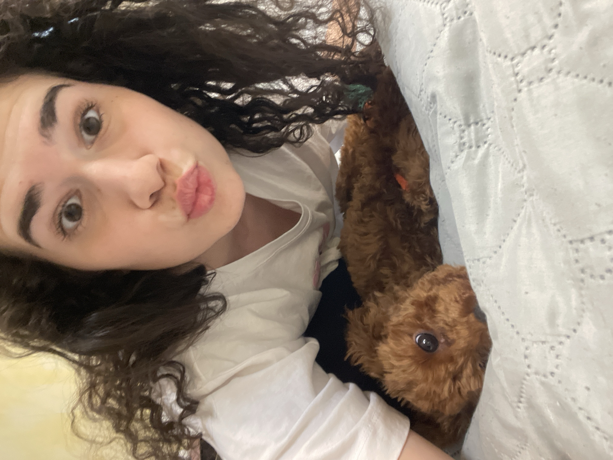 A young woman with curly dark hair and fair skin is taking a selfie indoors. She is wearing a white t-shirt. She has her lips puckered in a kiss expression. A brown poodle is partially concealed under a white textured blanket next to her. The location is not identifiable as Sofia, Bulgaria from the given visual information.