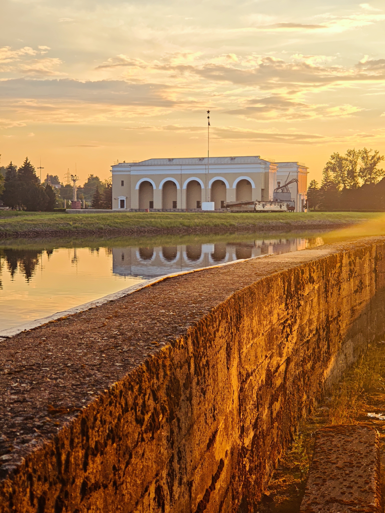 The image captures a sunrise scene at a waterfront, likely in the city of Vodnik SNT, Russia. In the foreground, a weathered stone wall, illuminated by the warm, golden light of the rising sun, runs diagonally across the frame. The wall's texture is rough, with visible moss and irregularities. Beyond the wall, a calm body of water reflects the sky and the structures on the opposite bank. The water's surface is smooth, with subtle ripples. Across the water, a long, light-colored building with a series of arched openings stands prominently. This building is set against a backdrop of greenery, including trees and well-maintained grass. The sky above is painted with soft, warm hues of orange and yellow, with scattered clouds catching the sunlight. Electrical poles and wires are visible in the distance, suggesting the presence of infrastructure. A small flag is atop a pole in front of the building. The overall atmosphere is serene and tranquil, bathed in the soft glow of dawn. There are no people or animals visible in the frame.