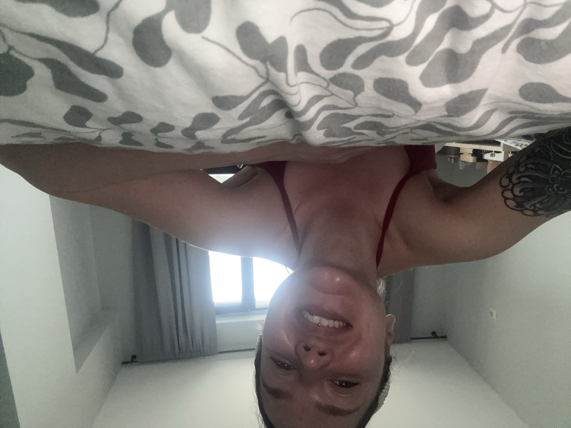 An indoor, upside-down selfie captures a person, likely a woman, smiling brightly at the camera. Her head and upper torso are visible, with her face showing a cheerful expression and bared teeth. She has a philtrum piercing and a prominent, detailed dark tattoo on her right arm. She is wearing a red top or swimsuit strap.
Above the subject, a white blanket or duvet with a repeating grey, organic, leaf-like pattern dominates the upper portion of the frame, partially obscuring the view. In the background, behind the subject's head, a large window allows bright natural light into the room, suggesting it is daytime. Grey curtains are visible on either side of the window, and the walls are light-colored. The setting appears to be a modern interior space, potentially a bedroom. No text is visible in the image. The location context provided for the image is Burgas, Bulgaria.