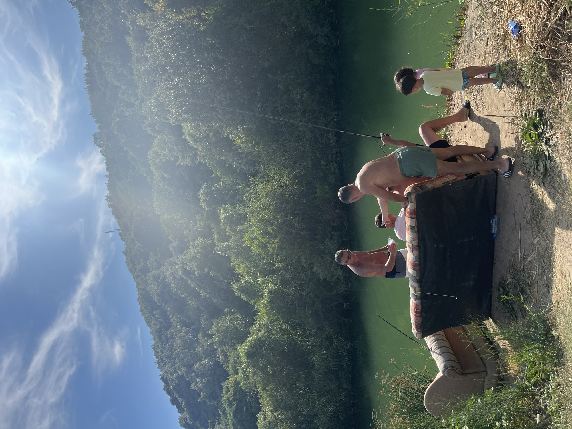 The image depicts a sunny day scene near a body of green water, possibly a lake or river, in Prijevor, Montenegro. A lush, forested hill rises in the background under a bright blue sky with scattered clouds. In the foreground, two sofas are placed on a dirt bank next to the water. Several individuals are present.
One man, shirtless and wearing swim trunks, stands near the water's edge, holding a clear container. Another shirtless man in green shorts is seated on one of the sofas, holding a fishing rod. A third person, wearing a pink shirt, is seated on the other sofa. Two young children, a boy and a girl, stand near the sofas. One of the fishing rods is cast into the water. There is a plastic bottle on the ground near the sofas, and some dry vegetation and grass are visible around the seating. The overall impression is of a casual outdoor gathering with fishing and relaxation.