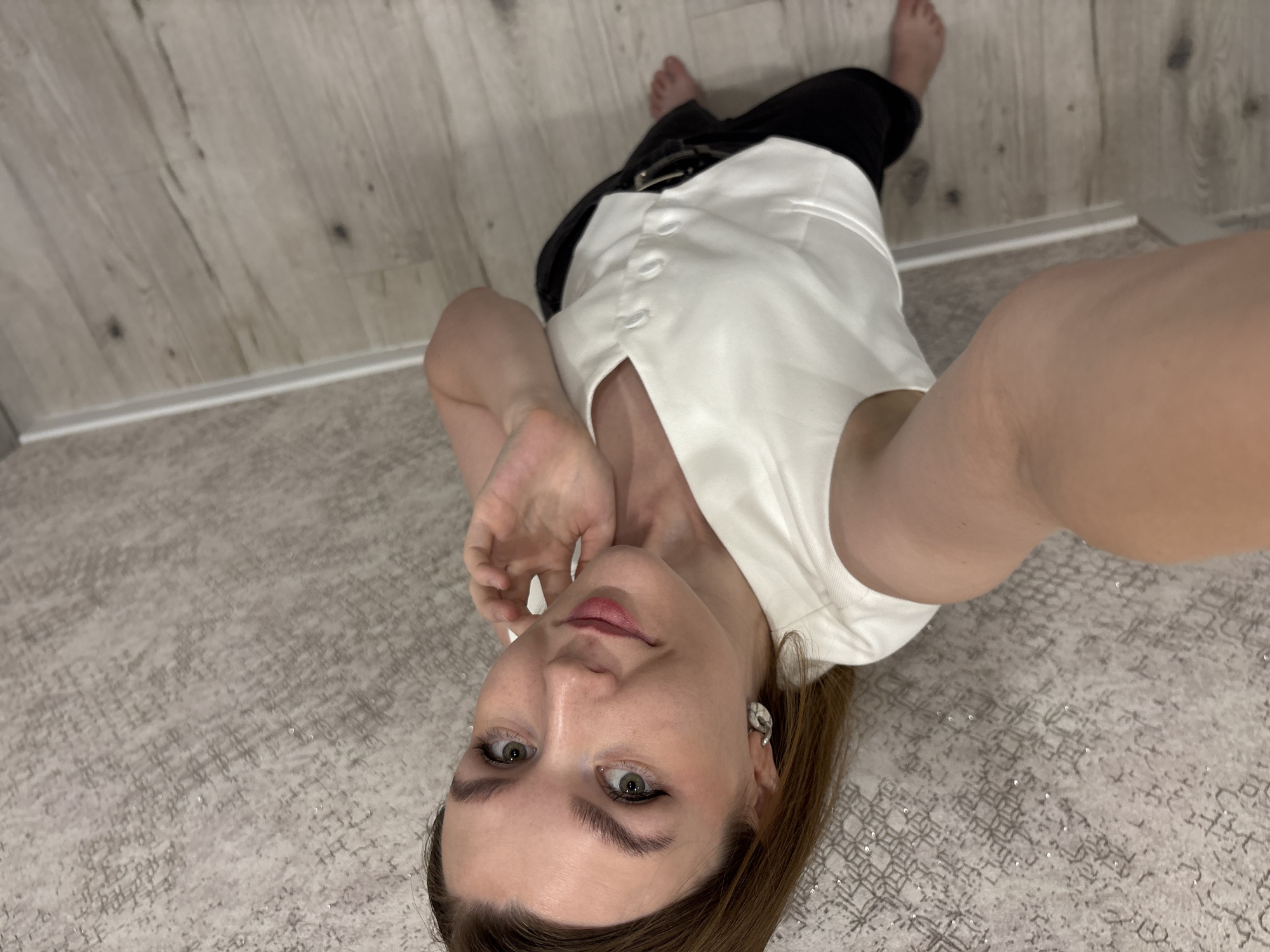 A woman is taking a selfie while lying on the floor. She is wearing a white vest over black pants with a belt. The floor has a patterned rug. The wall behind her is a light wood paneling. There is no indication that the photo was taken in Izhevsk, Russia.