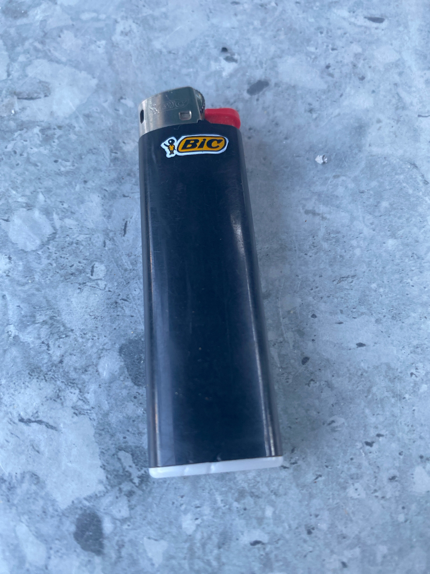 A close-up, overhead shot shows a black Bic lighter with a red ignition button resting on a textured, light grey surface that appears to be concrete. The lighter is positioned slightly off-center, with its top towards the left side of the frame. A faint "BIC" logo is visible on the top part of the lighter, near the metal spark mechanism. The concrete surface exhibits a mottled pattern of darker grey and white speckles, suggesting it might be an outdoor environment, possibly a sidewalk or patio. No individuals or other objects are present in the frame. The lighting indicates it is daytime, casting minimal shadows. The specific location within Novorossiysk, Russia, cannot be determined from the visual information provided.