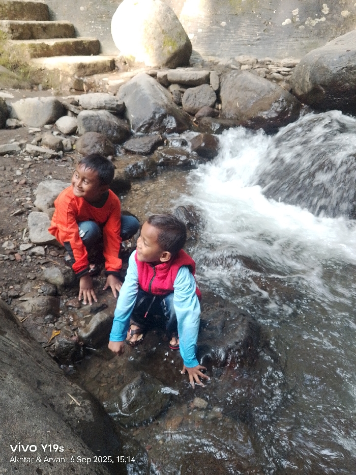 The image depicts two young boys, identified as Akthar and Arvan, interacting with a natural stream environment in Sukadana, Indonesia, captured at 15:14 on September 6, 2025, by a vivo Y19s device.
The boy on the left wears an orange long-sleeved shirt and dark trousers. He is in a kneeling or crouching position on the muddy bank of the stream, looking towards the left of the frame with a smiling expression. His hands are placed flat on the ground.
The boy on the right wears a red sleeveless vest over a light blue long-sleeved shirt, dark trousers, and open-toed sandals. He is also in a kneeling or crouching position, situated closer to the water's edge on a dark, wet rock, and appears to be looking in the same direction as the first boy. His hands are in contact with the wet surface of the rock or shallow water.
The environment is characterized by a rapidly flowing stream with visible whitewater, cascading over numerous dark, varied-sized rocks that form the stream bed and banks. The ground surrounding the stream is earthy and appears damp. In the upper left background, a set of rough, light-colored stone steps ascends, flanked by more rocks. A segment of a large, dark tree trunk or log is partially visible in the lower left foreground. The scene is illuminated by natural daylight.