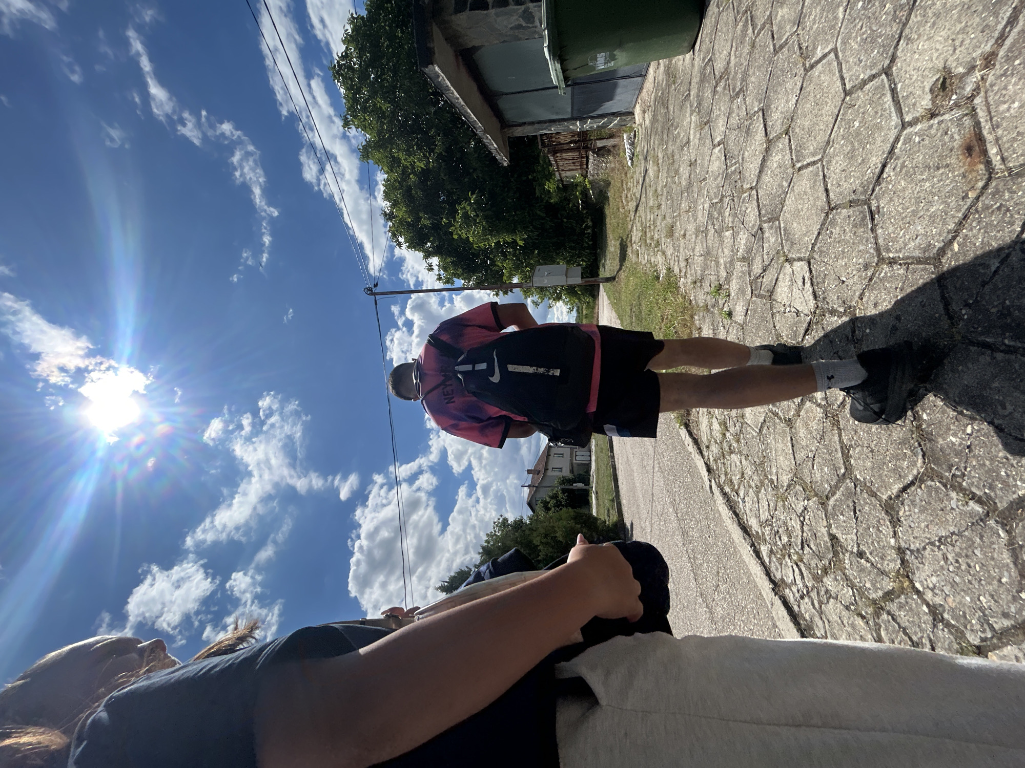 The image shows two people walking down a paved sidewalk. The person in the foreground is partially visible from the side, wearing a black t-shirt and grey sweatpants. The second person, in the mid-ground, is walking away from the camera, wearing a tie-dye t-shirt, black shorts, and a black backpack. The background features a blue sky with scattered clouds and the sun, trees, a building with a garage door, and a green trash can. The sidewalk is made of hexagonal concrete pavers. The scene appears to be in a residential area.